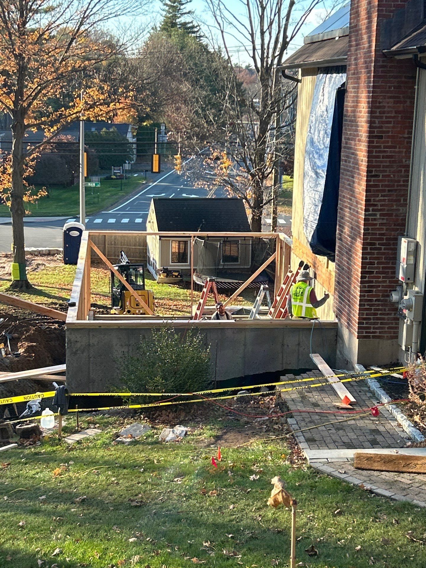 Construction site next to a brick building and road. A small wooden structure and heavy machinery are present. Yellow caution tape.