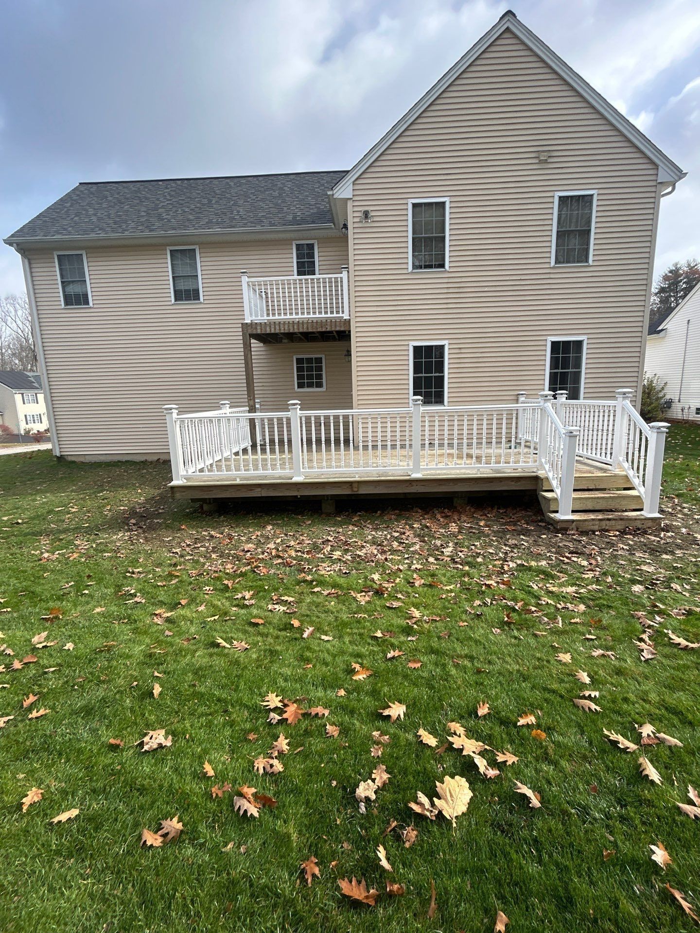 Beige two-story house with a wooden deck. Deck has white railing, steps leading to the yard, and is covered in fallen leaves.