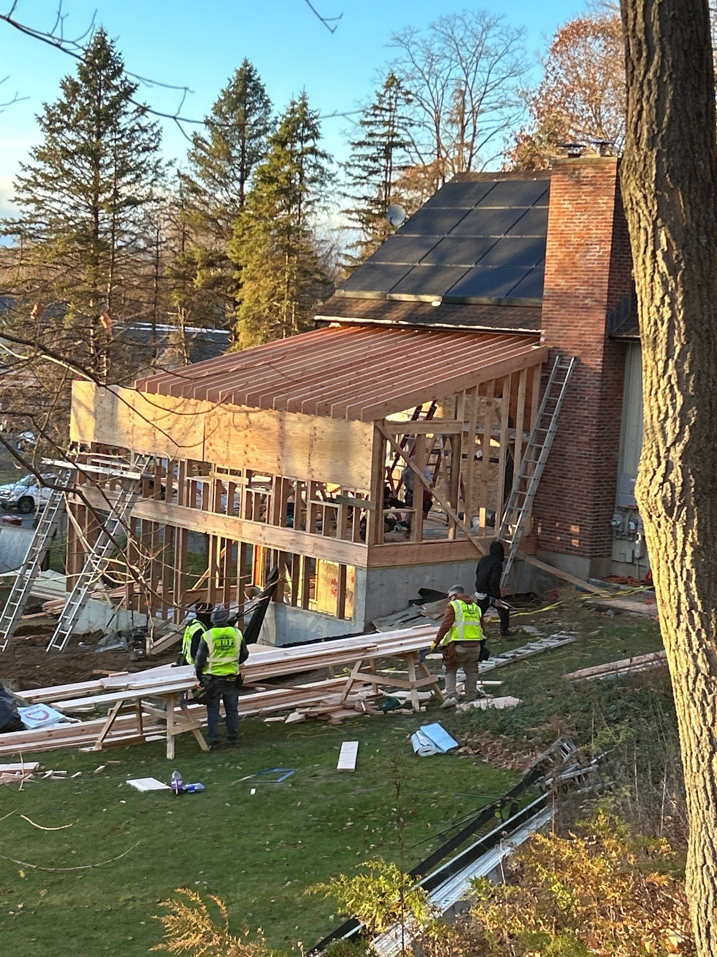 Construction workers build an addition to a brick house. Wooden frame, copper-colored roof, and ladders are visible.
