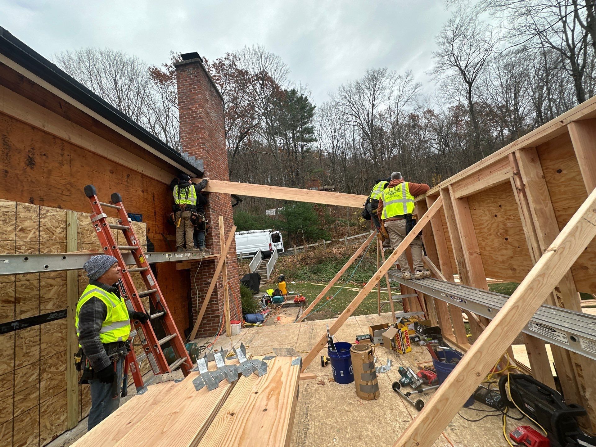 Construction workers building a wooden frame. They wear vests, work outdoors with tools, and near a brick chimney.