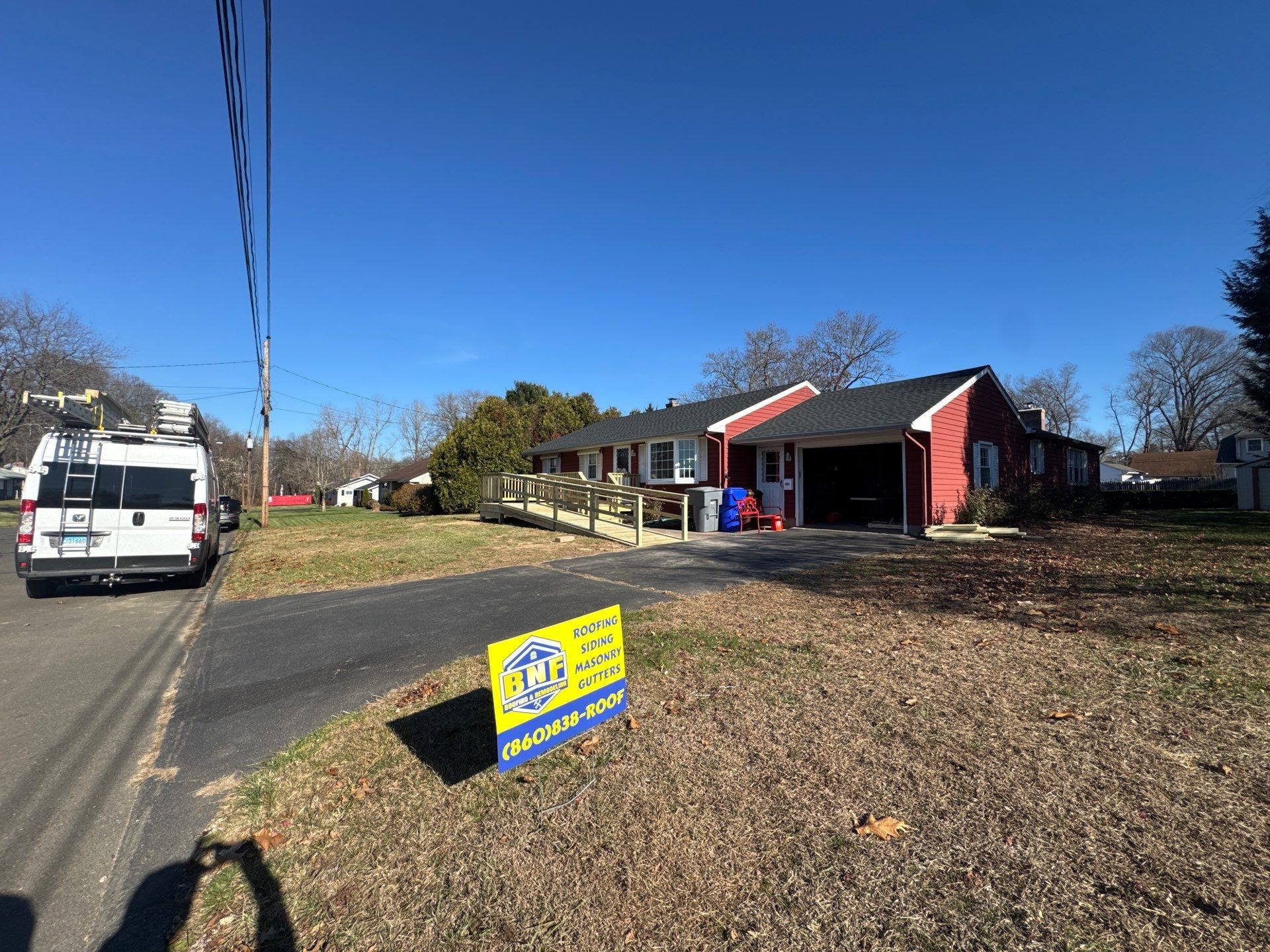 House with red siding and black roof; sign in front indicates roofing services; van parked nearby.