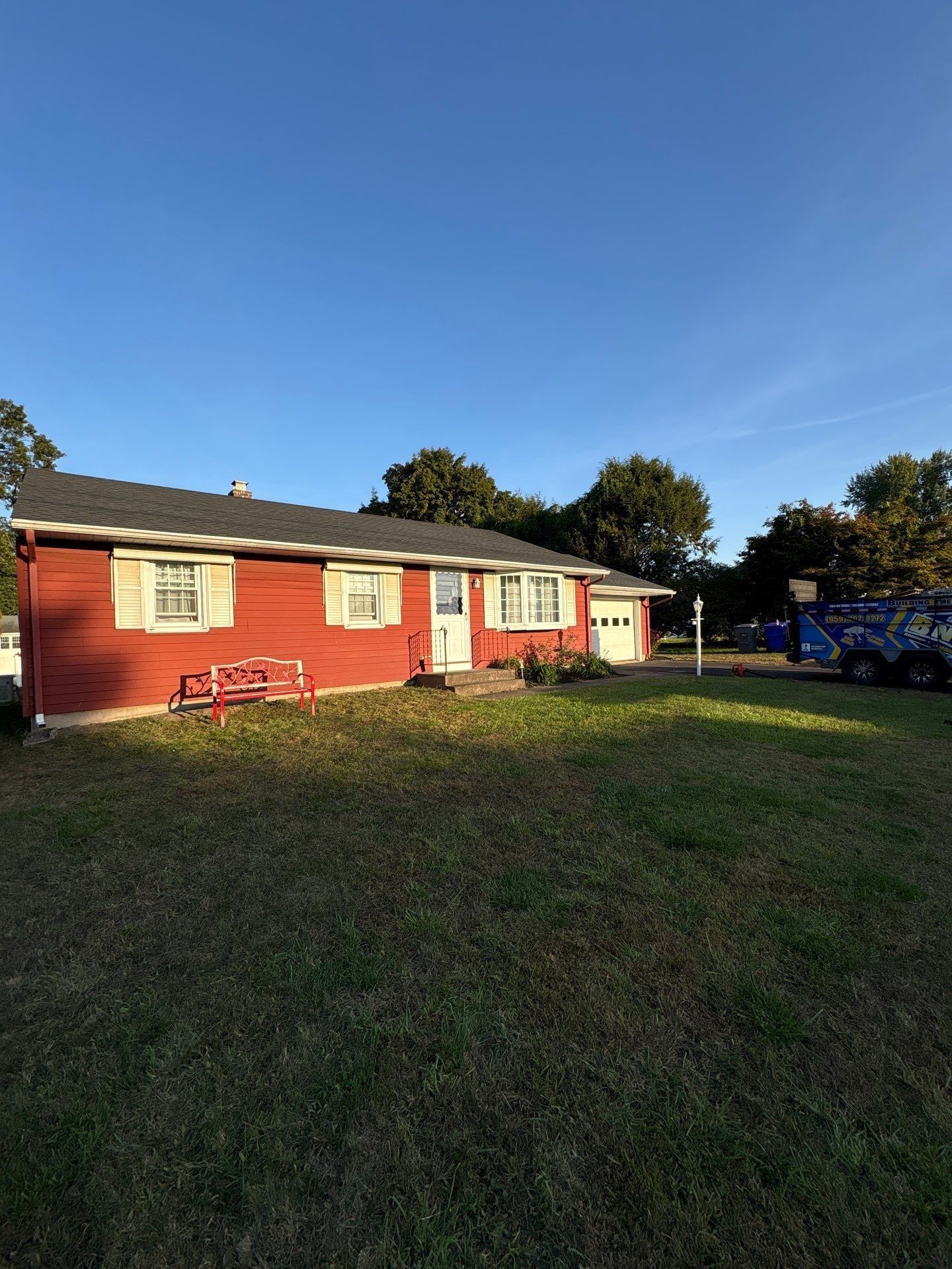 Red one-story house with tan shutters and a garage on a grassy lot under a blue sky.