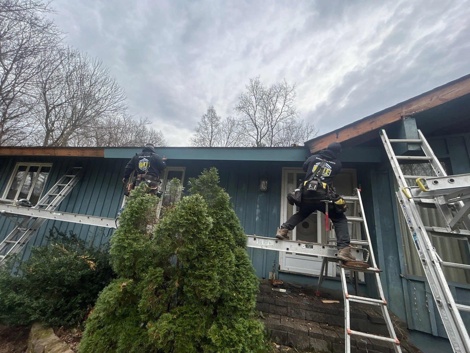 Two workers on ladders repairing the blue siding of a house, cloudy sky above.