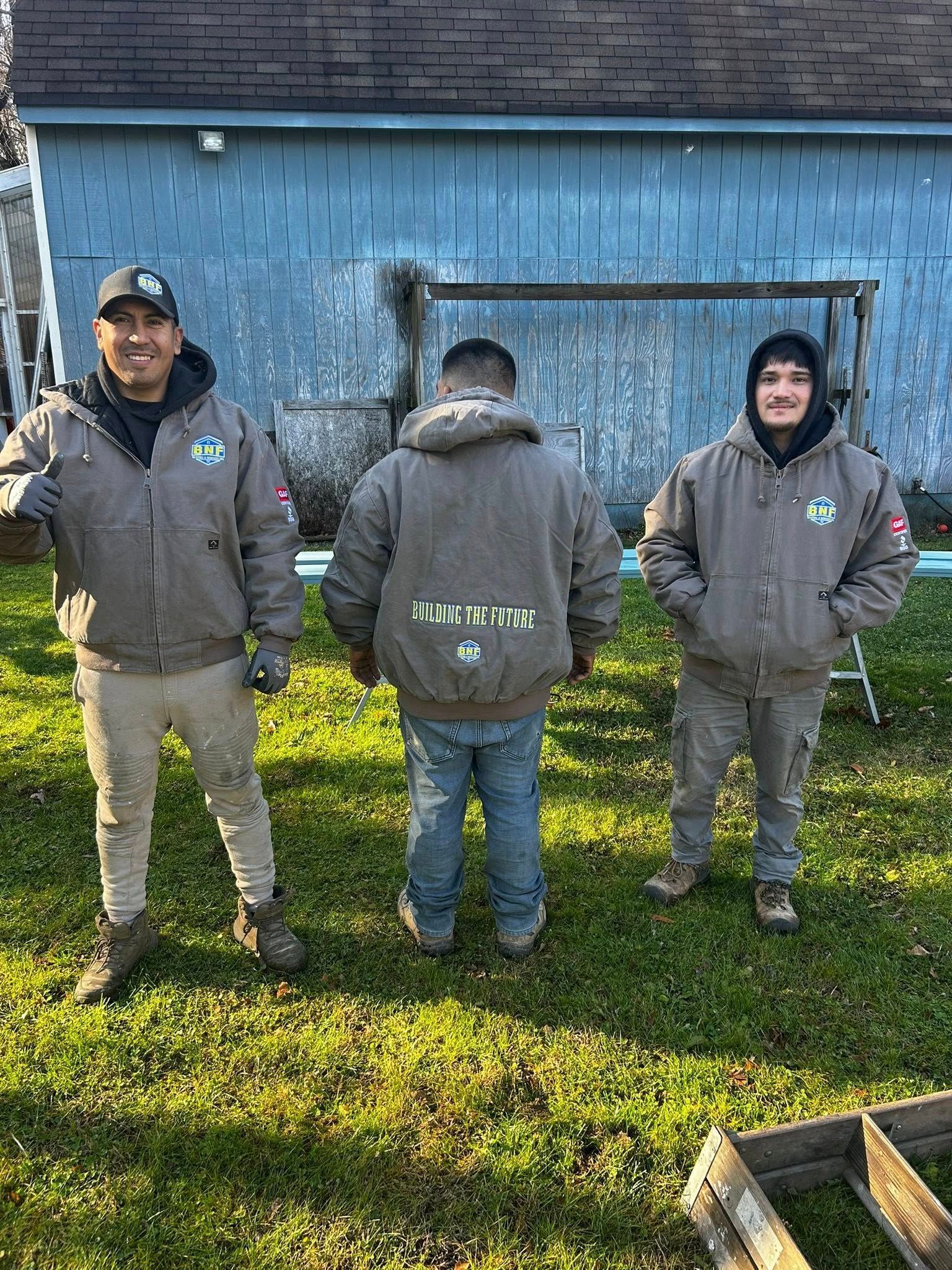 Three people wearing jackets in front of a blue building, standing on grass. One is facing the camera.