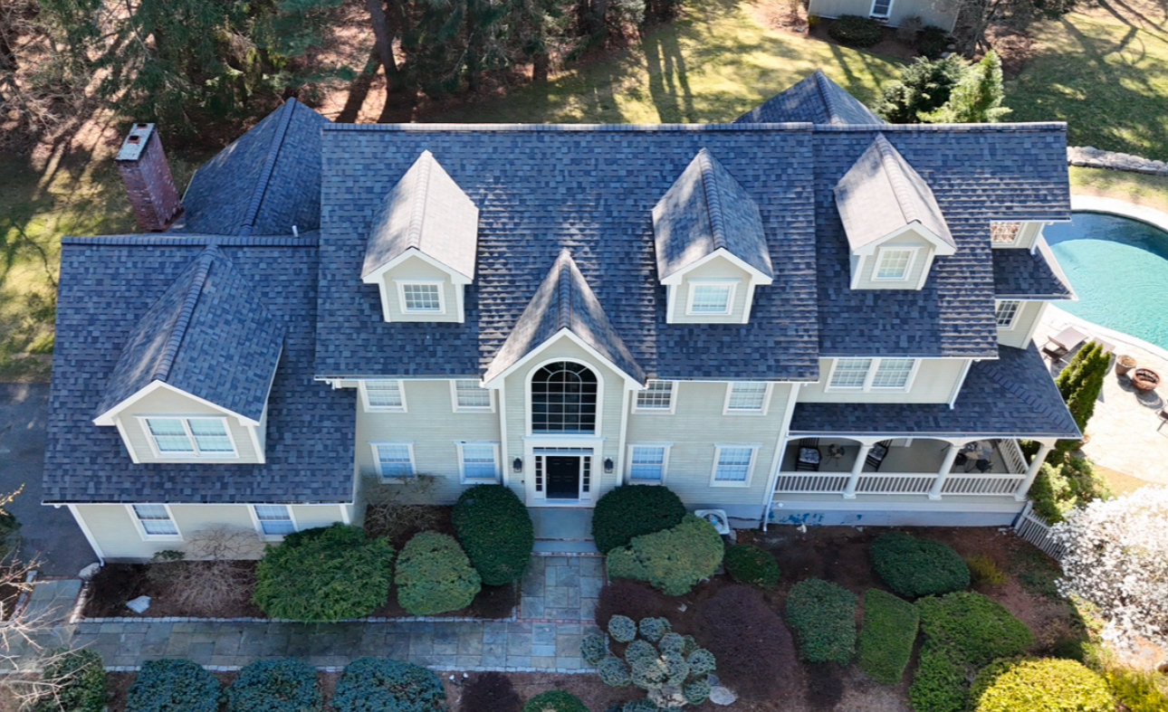Large two-story house with blue roof, dormers, and a swimming pool in the background.