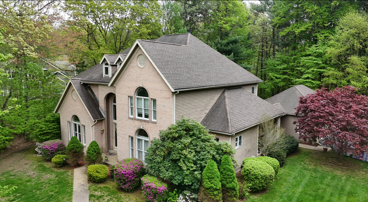 House with tan brick exterior, arched windows, and a dark shingled roof, surrounded by green trees and shrubs.