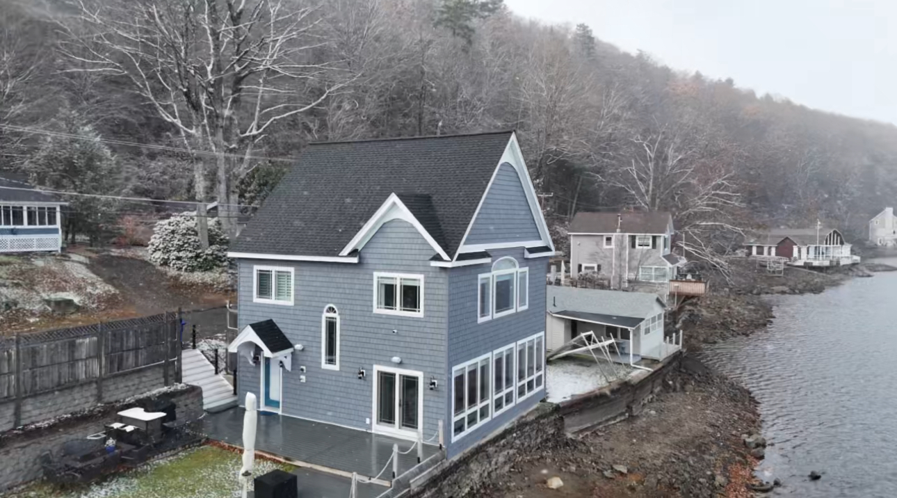 Two-story gray house on a waterfront with snow on the ground; trees in the background.