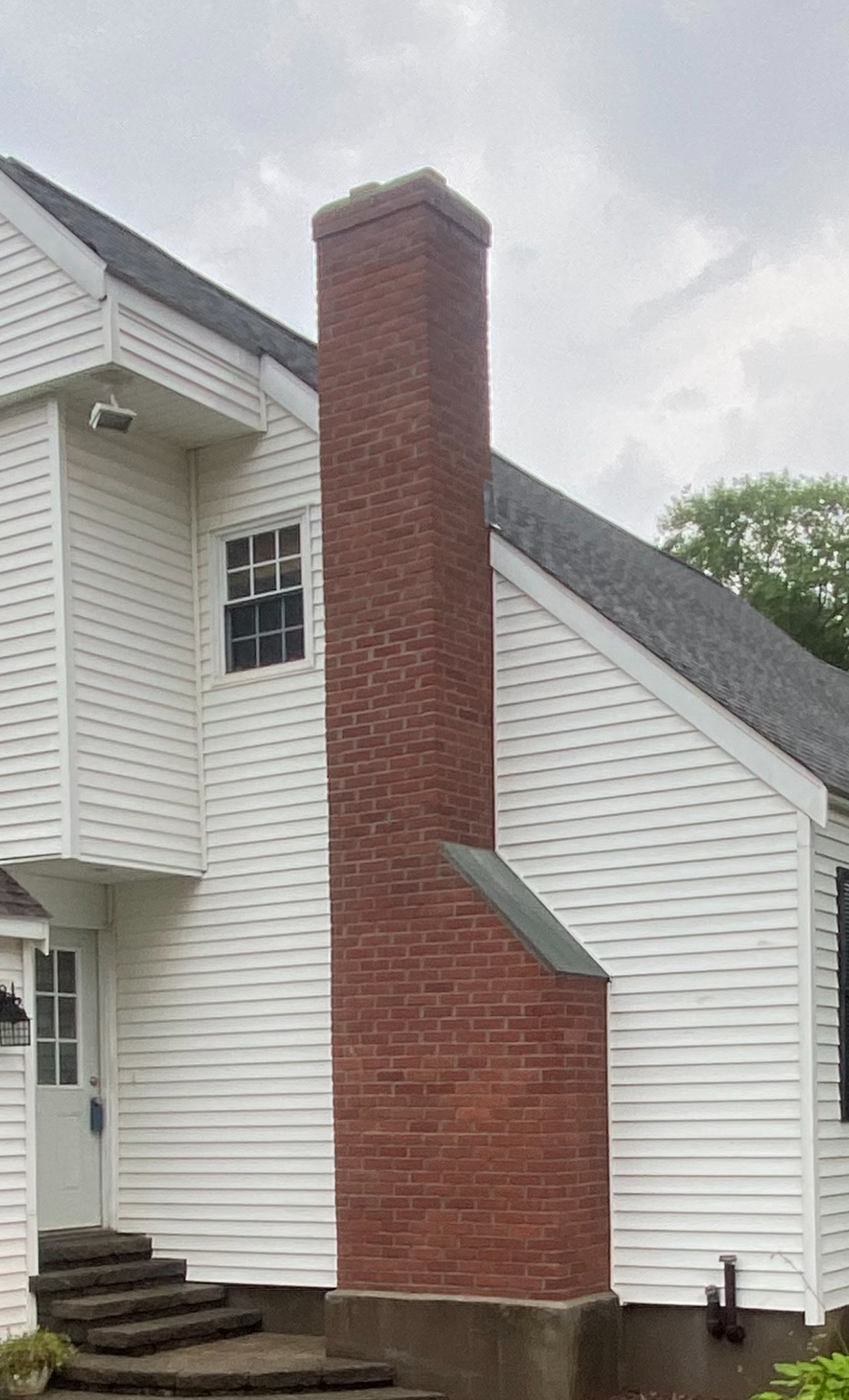 Brick chimney attached to a white-sided house with a sloping roof and a set of stairs.