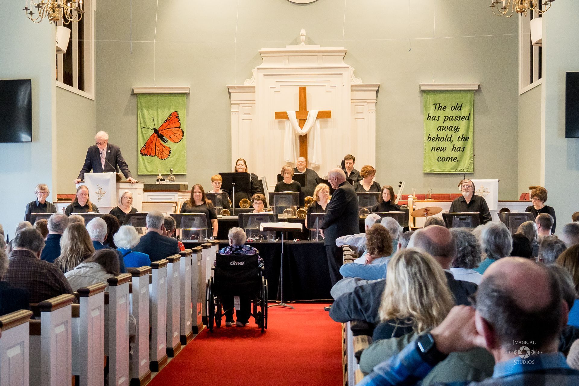 Church service with handbell choir and congregation. People in pews, red carpet, butterfly banner, cross.