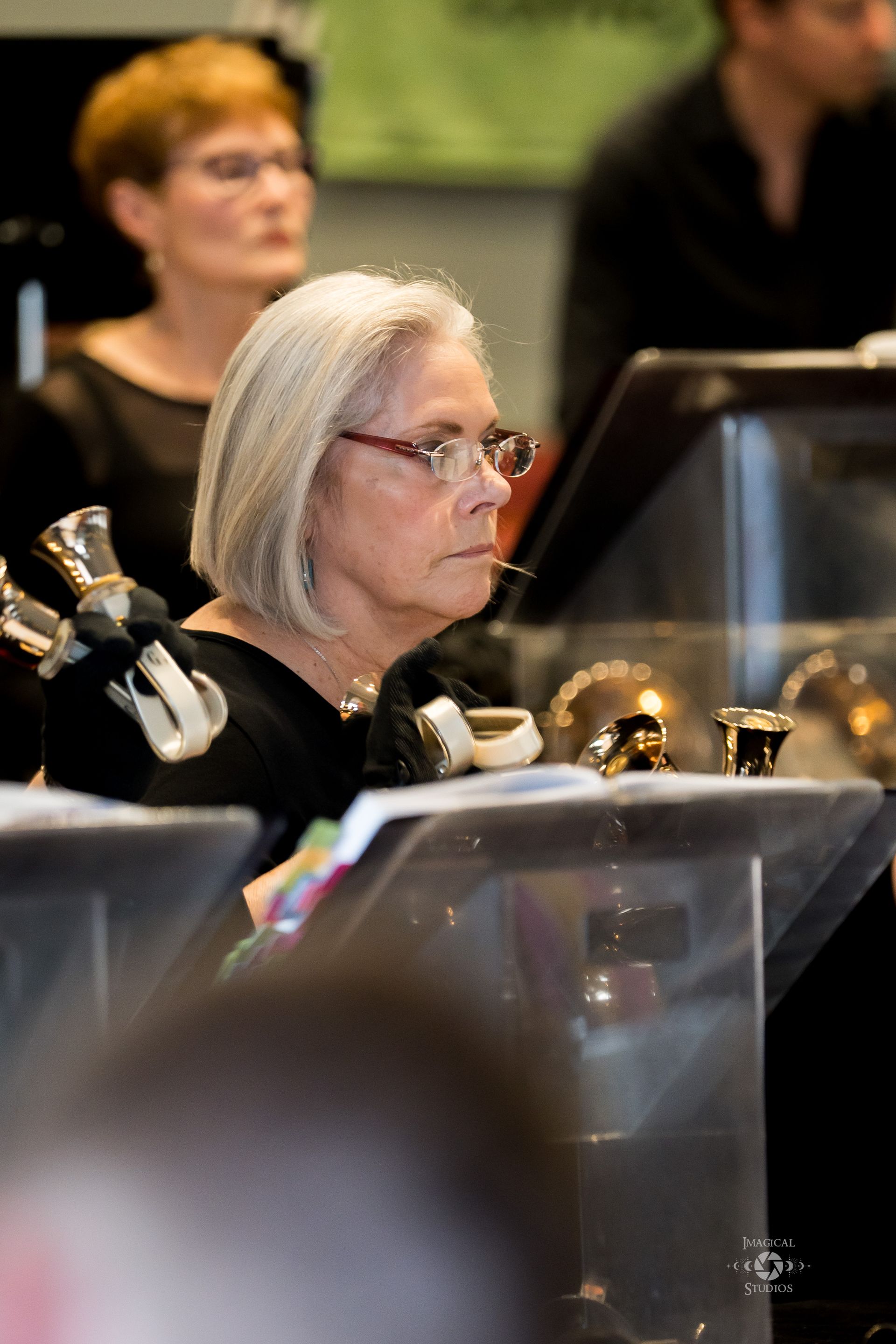 Woman with glasses in a handbell ensemble, looking at sheet music. Setting appears to be a concert hall.