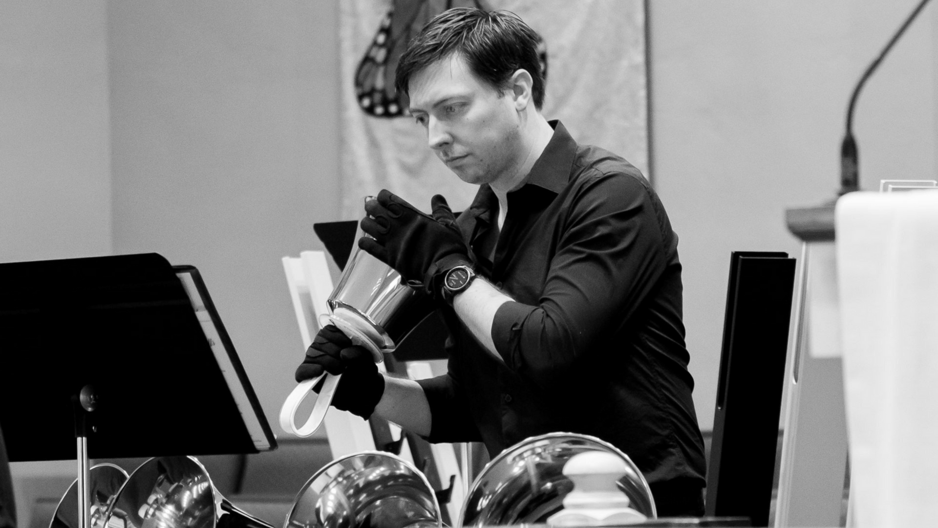 Man in black shirt, ringing handbells; music stand in foreground.