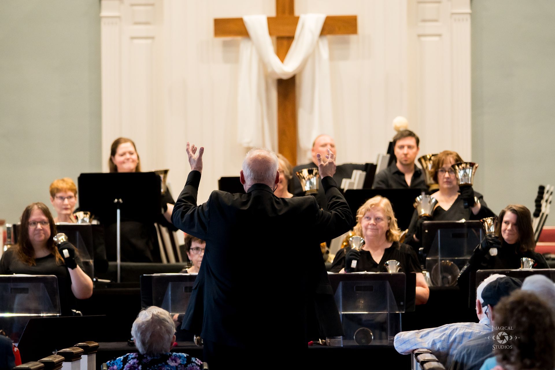 Handbell choir in church, conductor with arms raised, players behind music stands, cross in background.