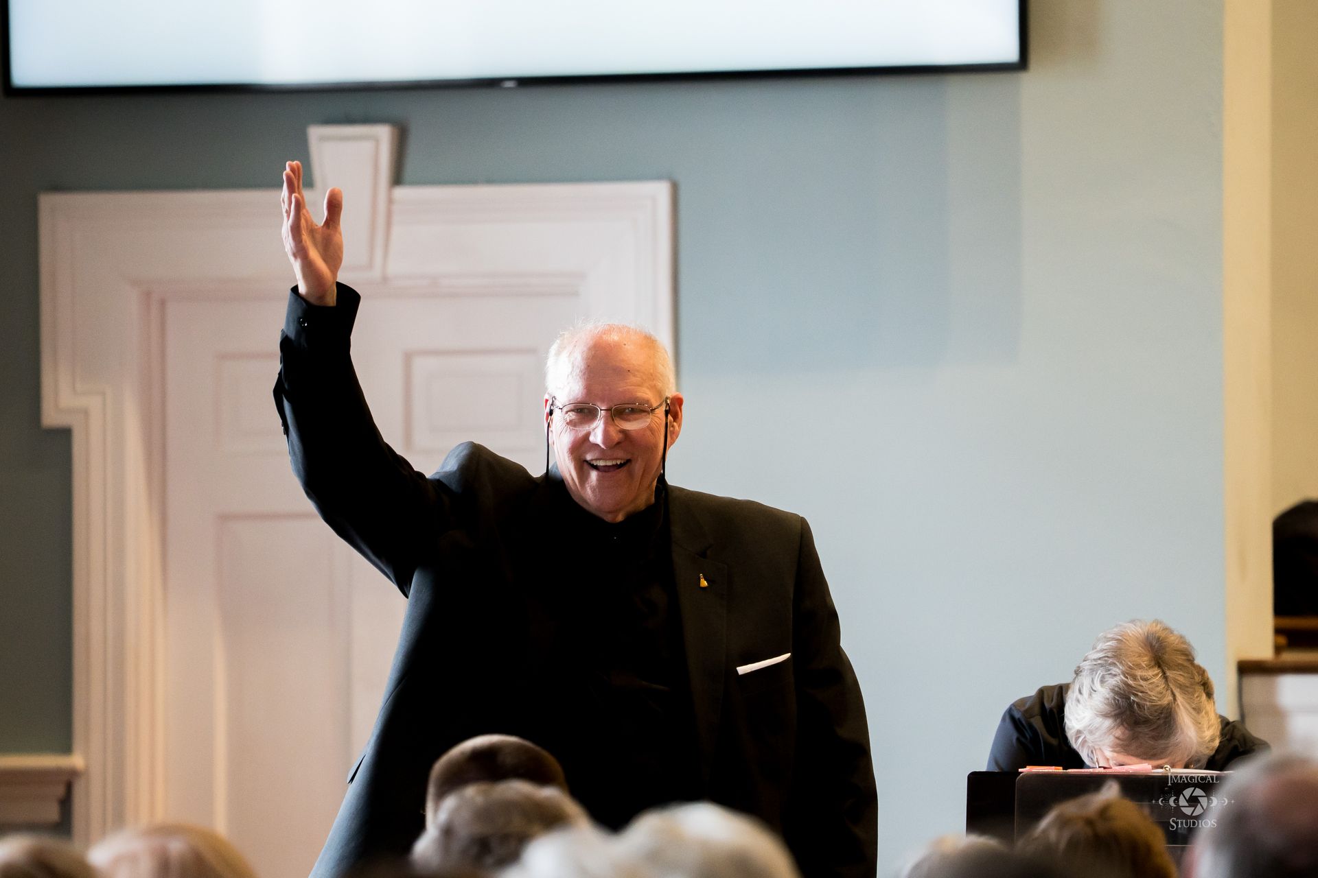 Man in black suit raises hand in a light-filled room; person in background bows head.