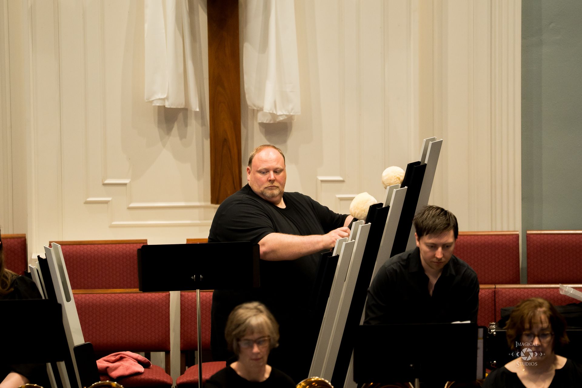 A man playing handbells in a church. Other musicians sit nearby, red chairs in the background.