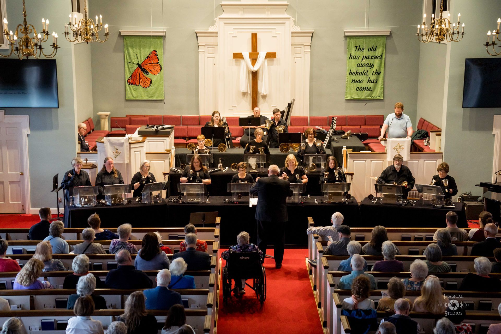 Handbell choir performing in church, conductor leads while audience listens.
