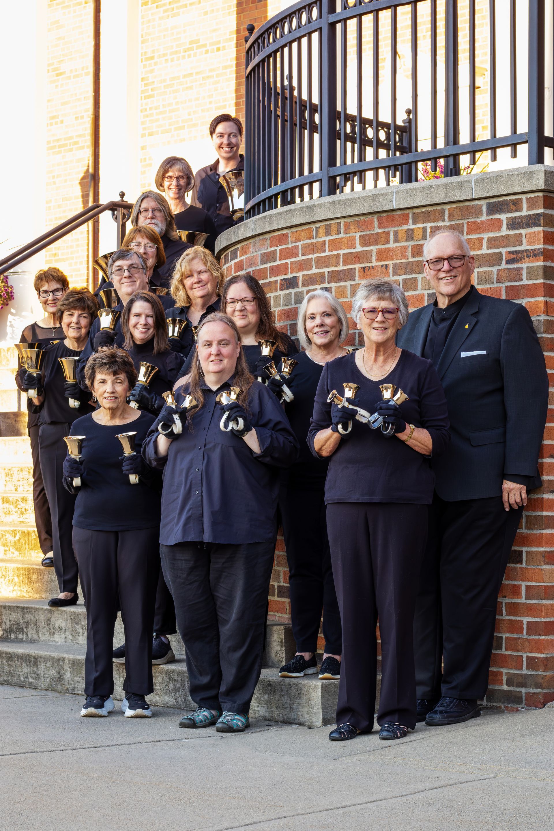 Group of people, smiling, holding bells. Standing on steps, near brick building.