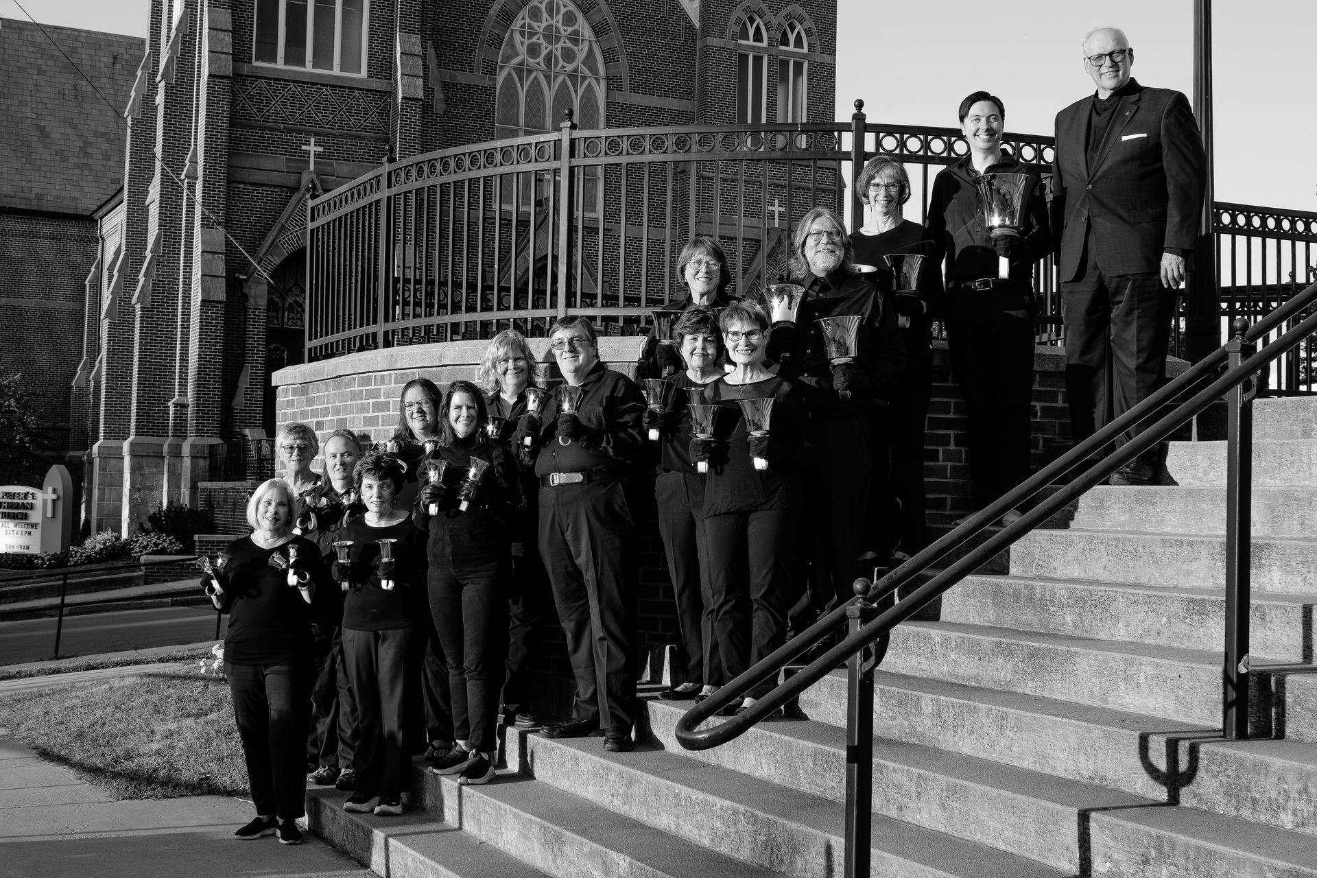 Choir group posing on church steps; people in dark attire holding handbells, man in suit, black and white.
