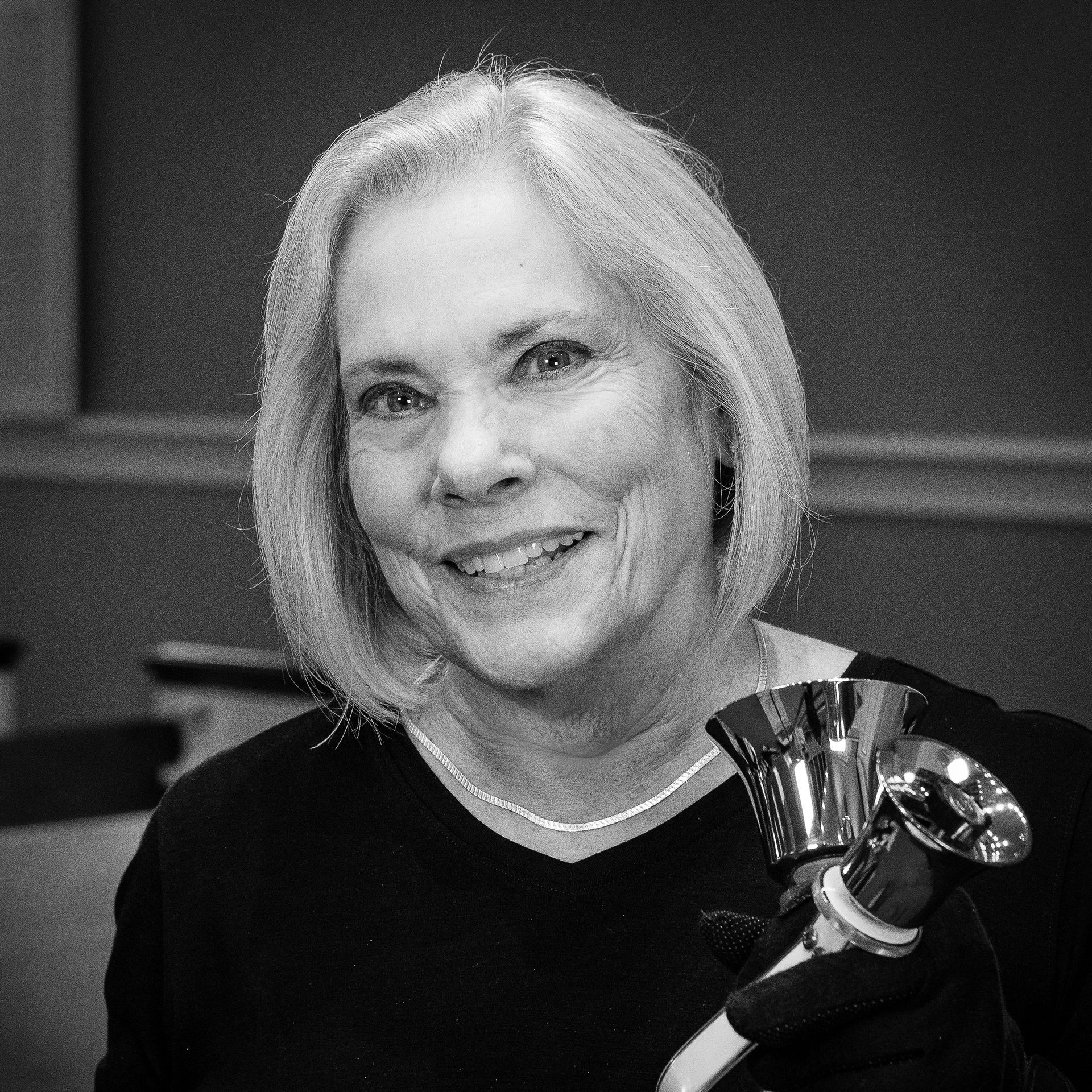 Woman with gray hair, smiling, holding a  handbell. Black top.