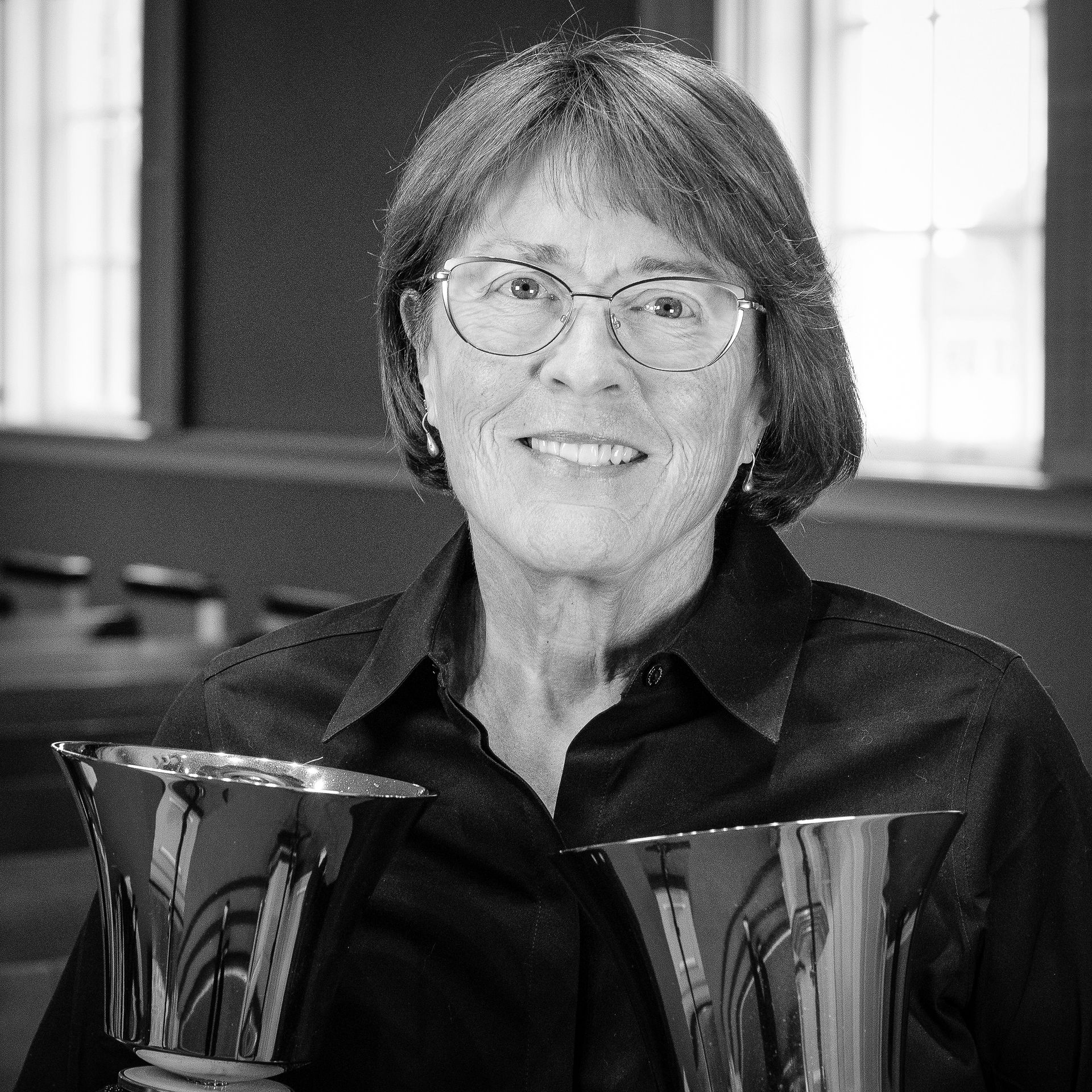 Woman in glasses smiling, holding two handbells.