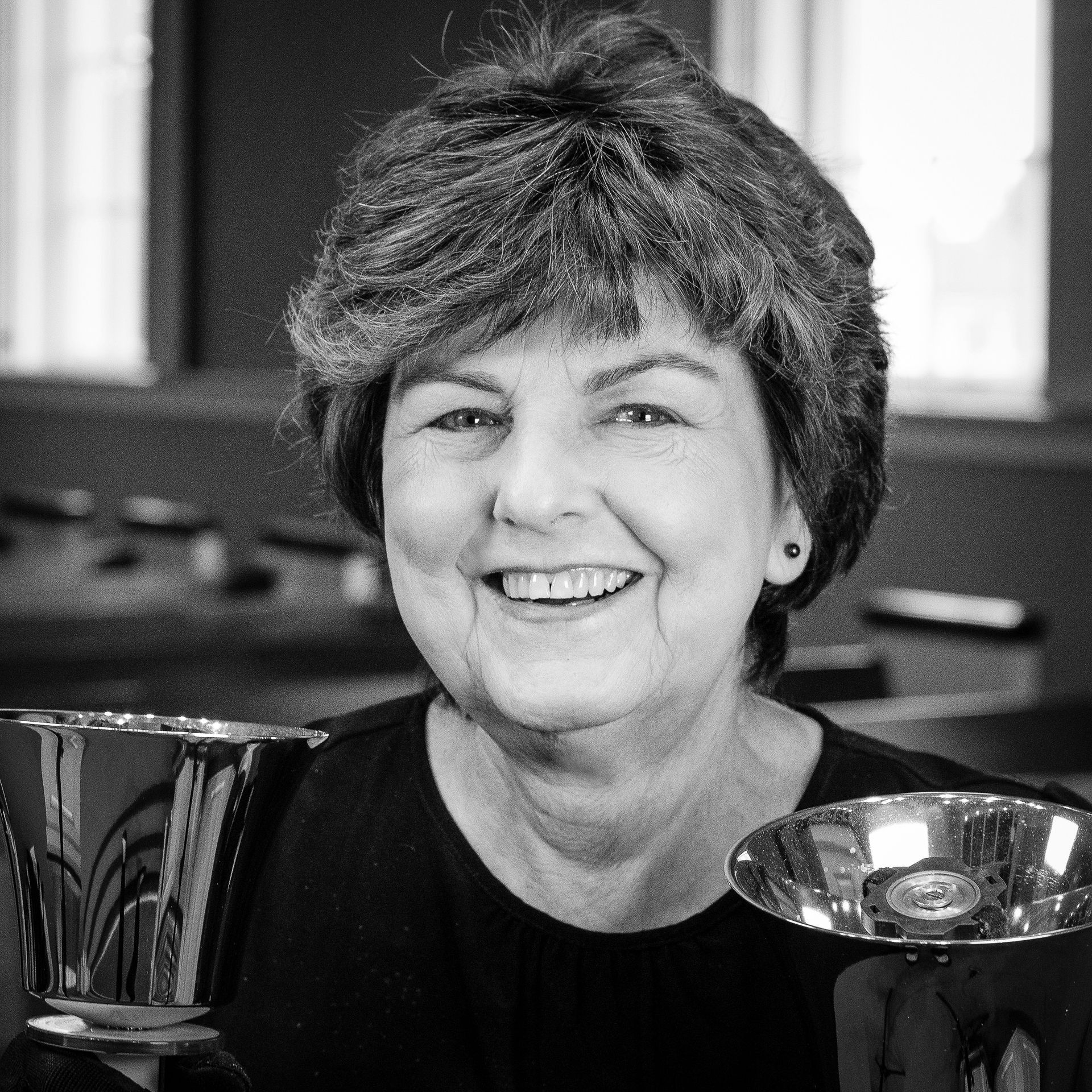 Woman smiling, holding two handbells, black top, indoor setting.