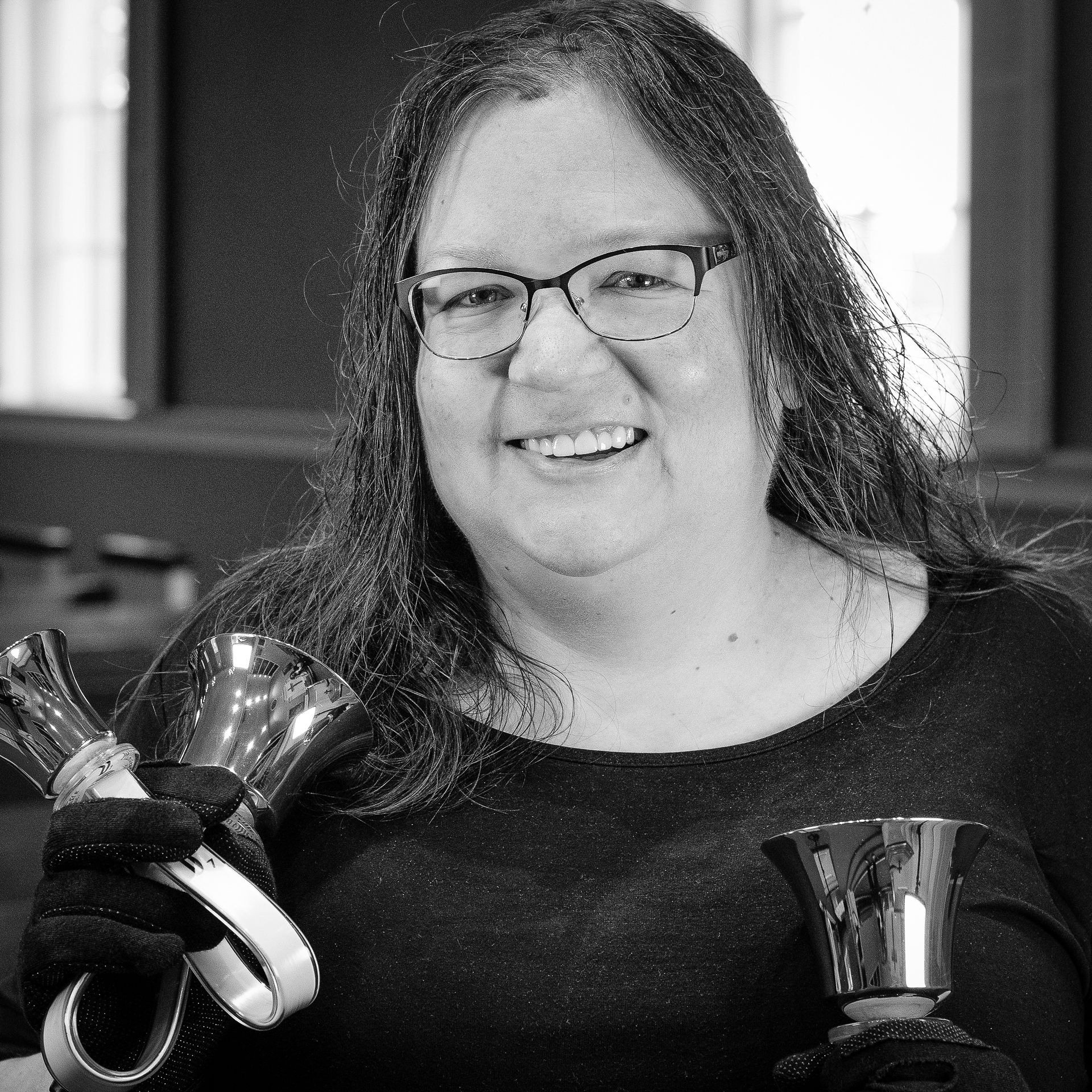 Woman with glasses and long hair holding handbells, smiling.