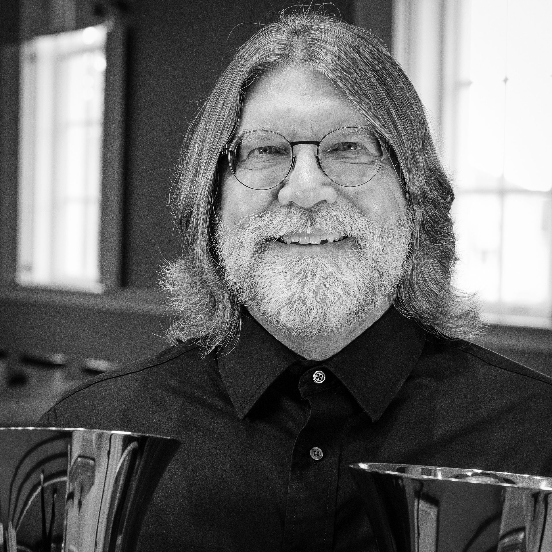 Man with long hair and beard, smiling, wearing glasses and a black shirt, holding handbells.