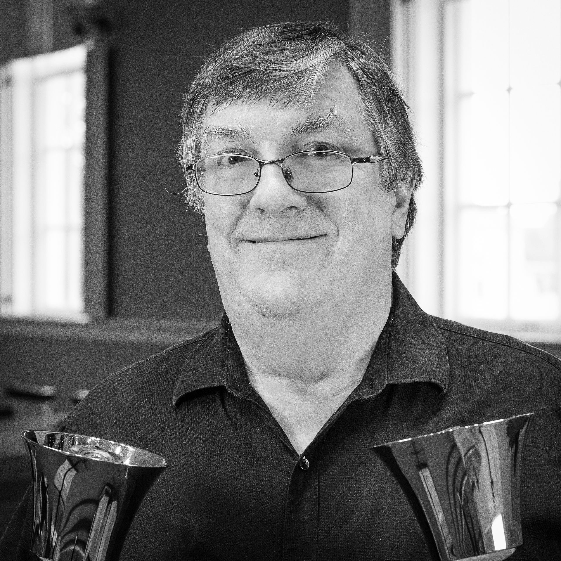 Man smiling, holding two handbells, in a room with windows.