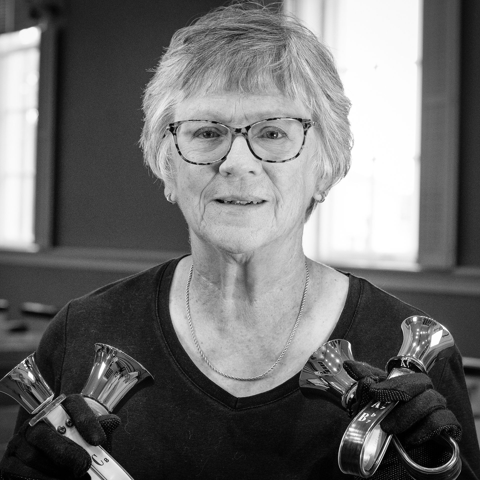 Older woman holding handbells, wearing glasses, smiling, indoor setting.