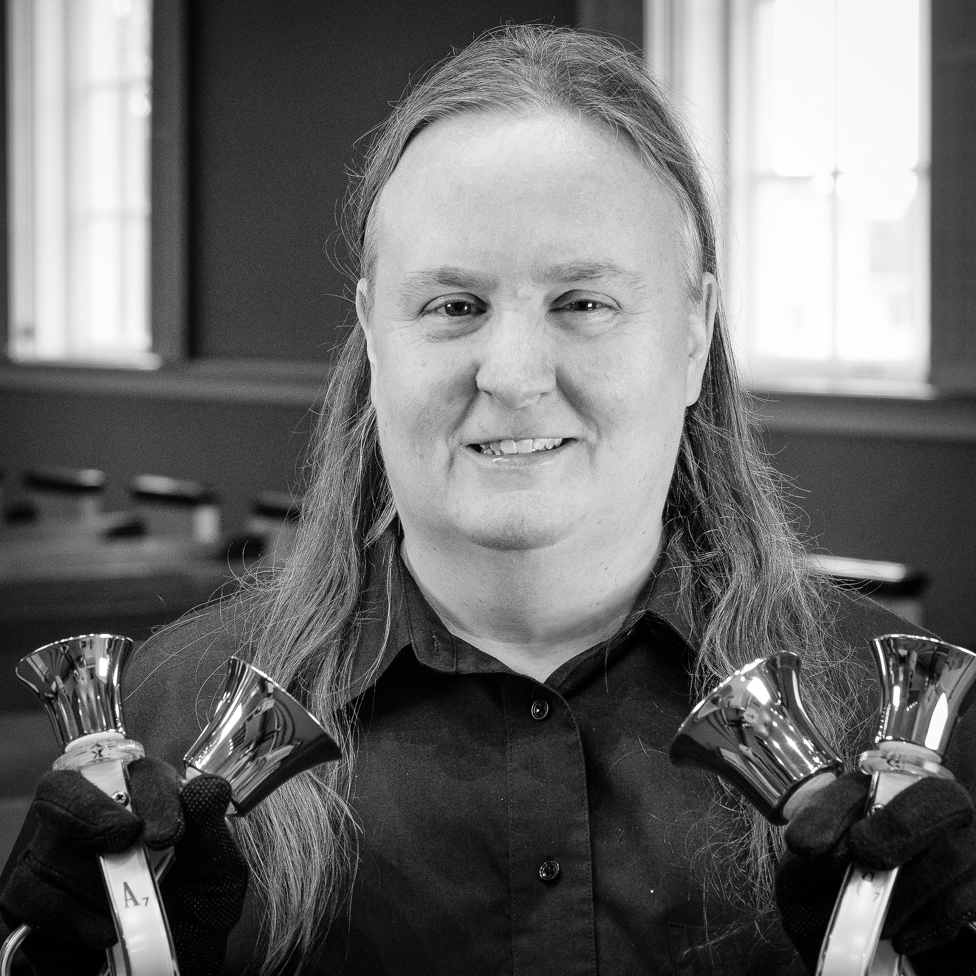 Woman with long hair, smiling, holding handbells, in a room with windows.