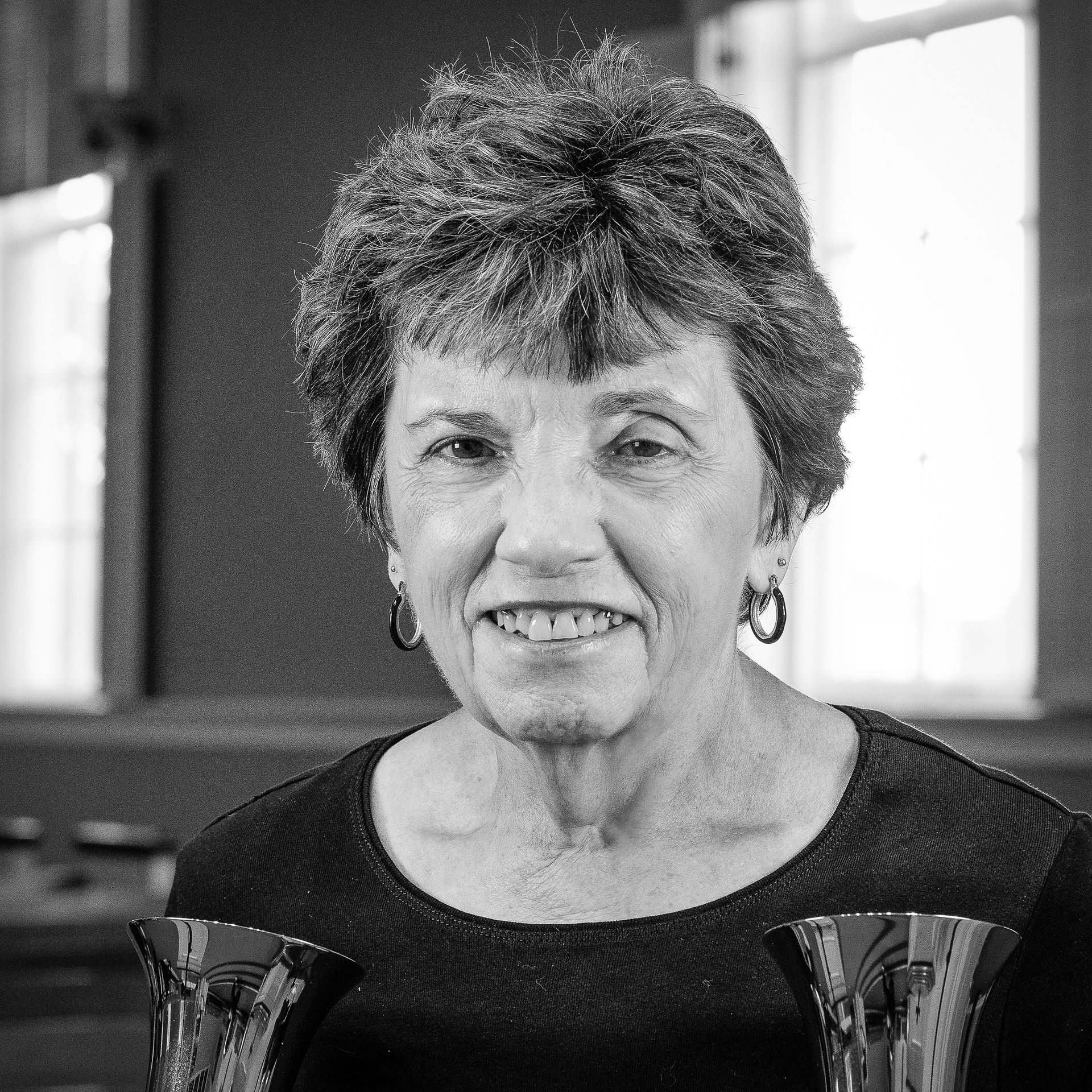 Smiling older woman with short, curly hair holds two handbells, indoors.