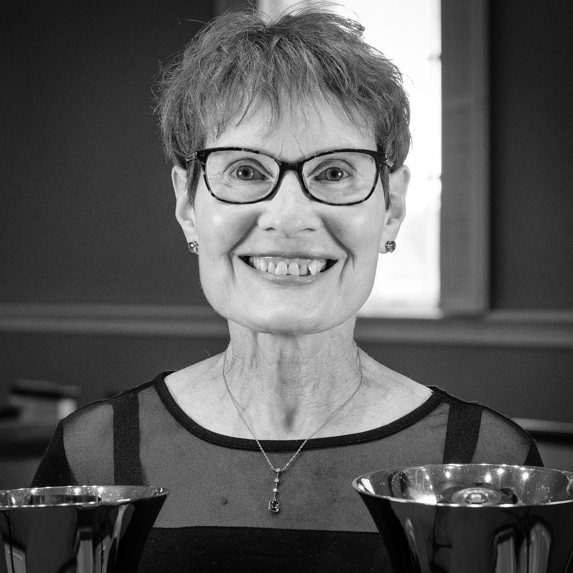 Woman with glasses smiling, holding handbells, inside a building.