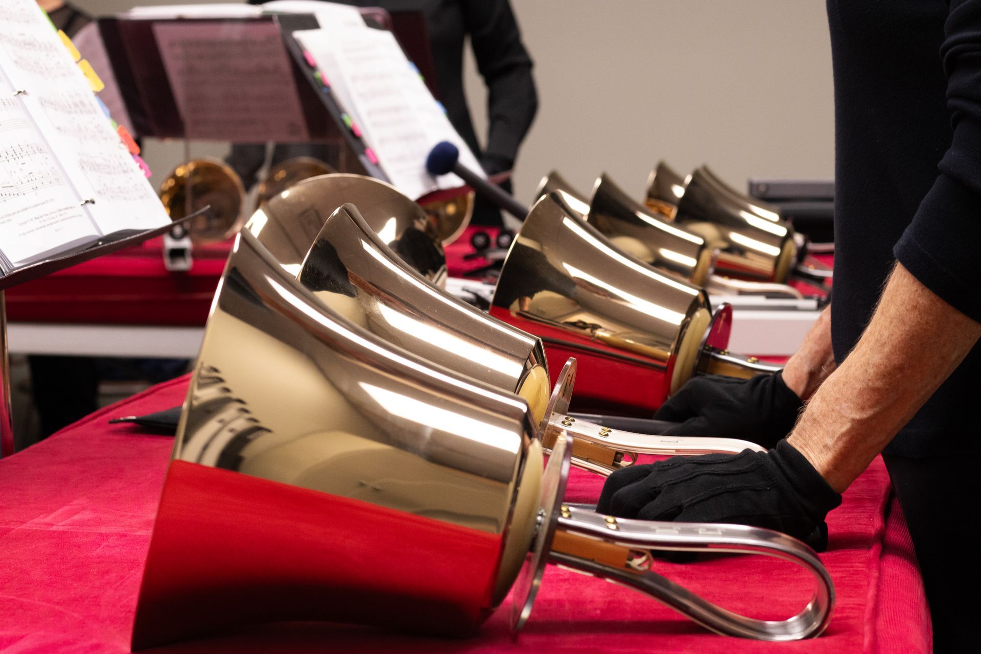 Hands playing handbells on a red table; music stand in background.
