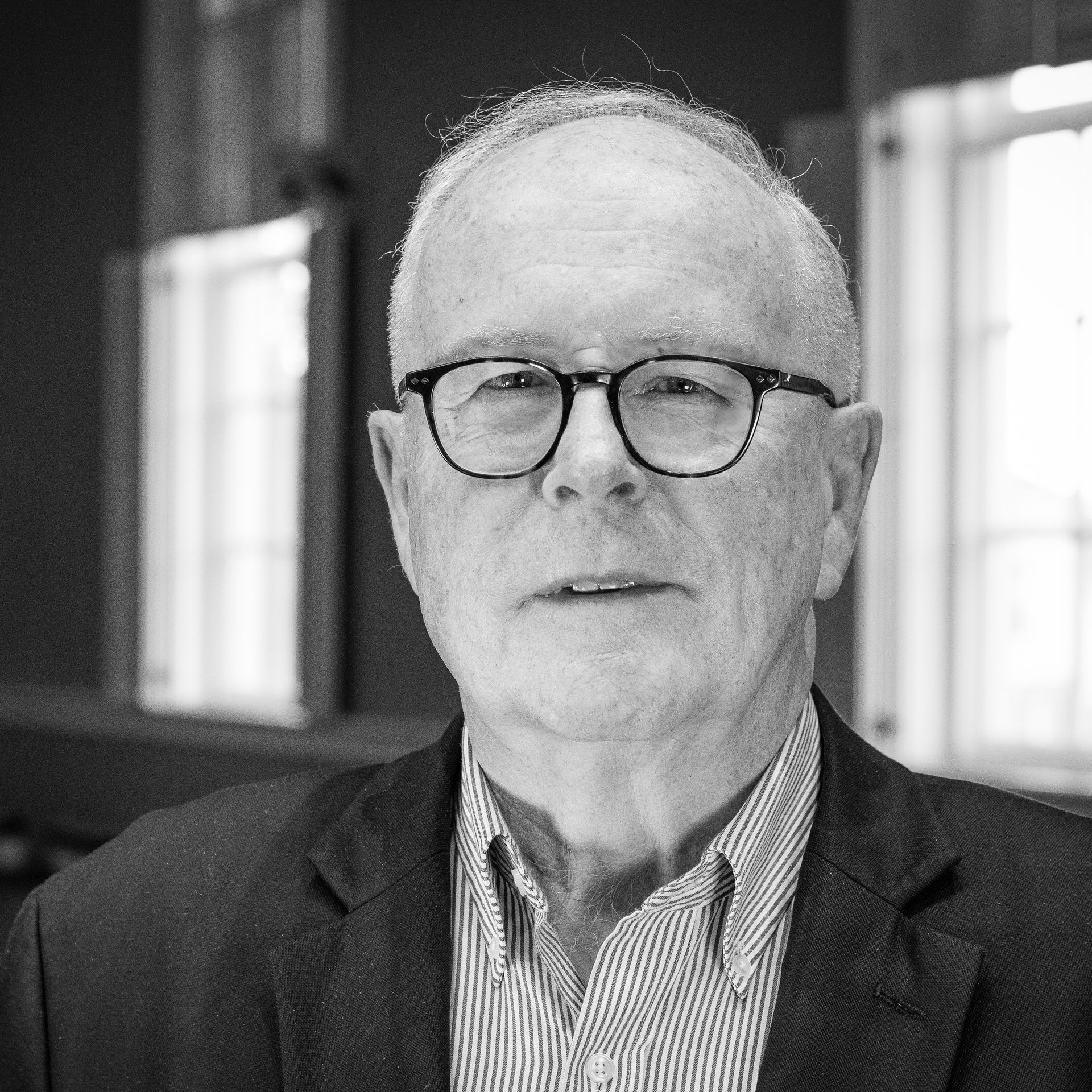 An older man with glasses wearing a blazer, striped shirt, in front of windows.