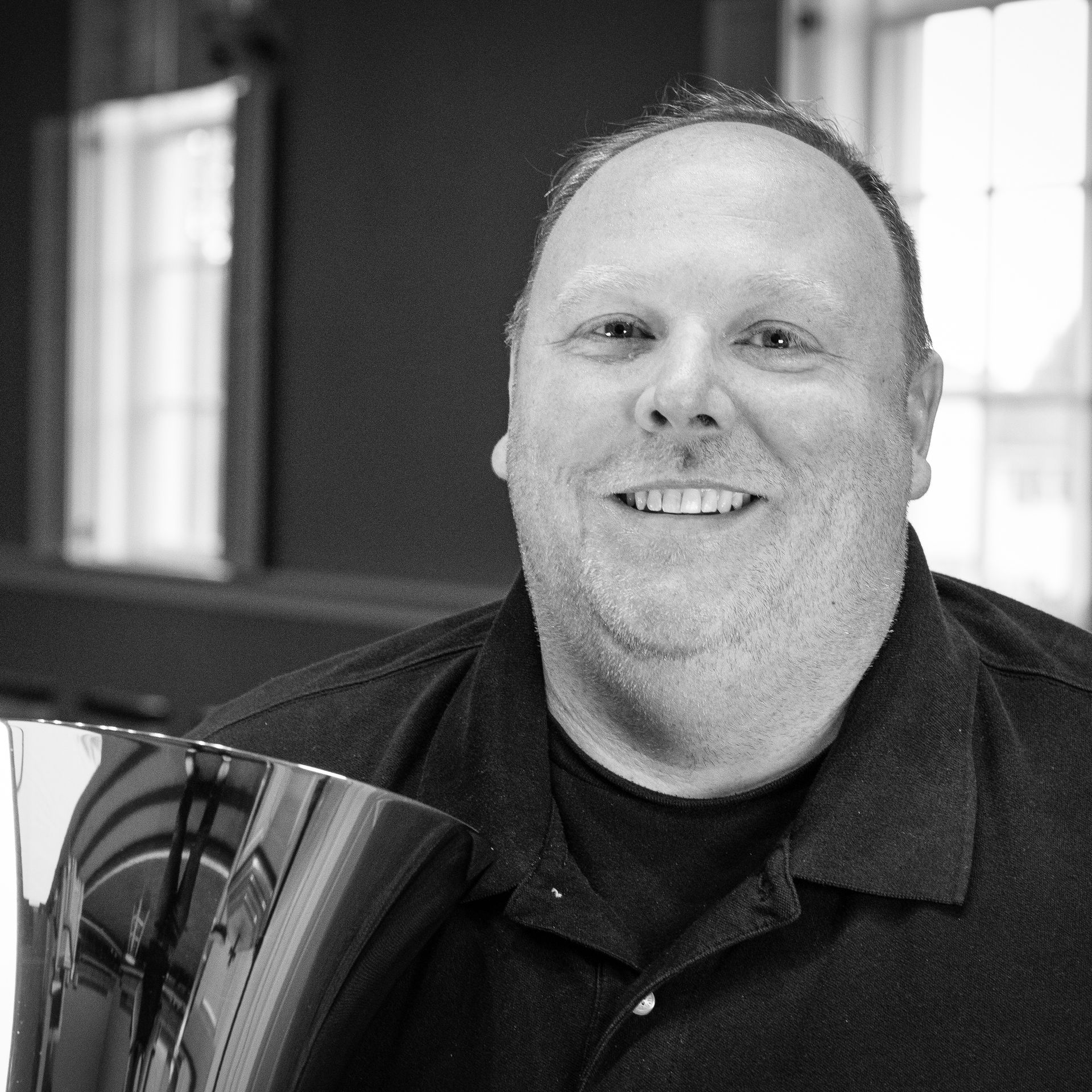 Man with a slight smile, holding a shiny handbell. Inside a building, window in background.