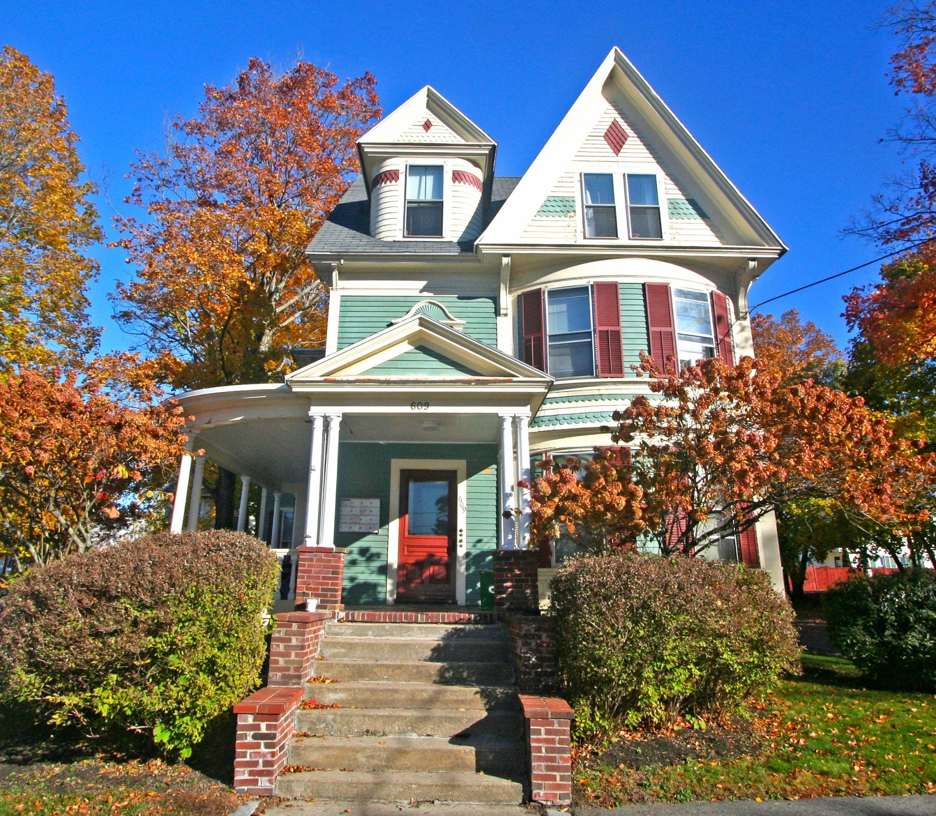 Exterior view of three story beige Victorian style home with red door, green panels, stairs to front porch.
