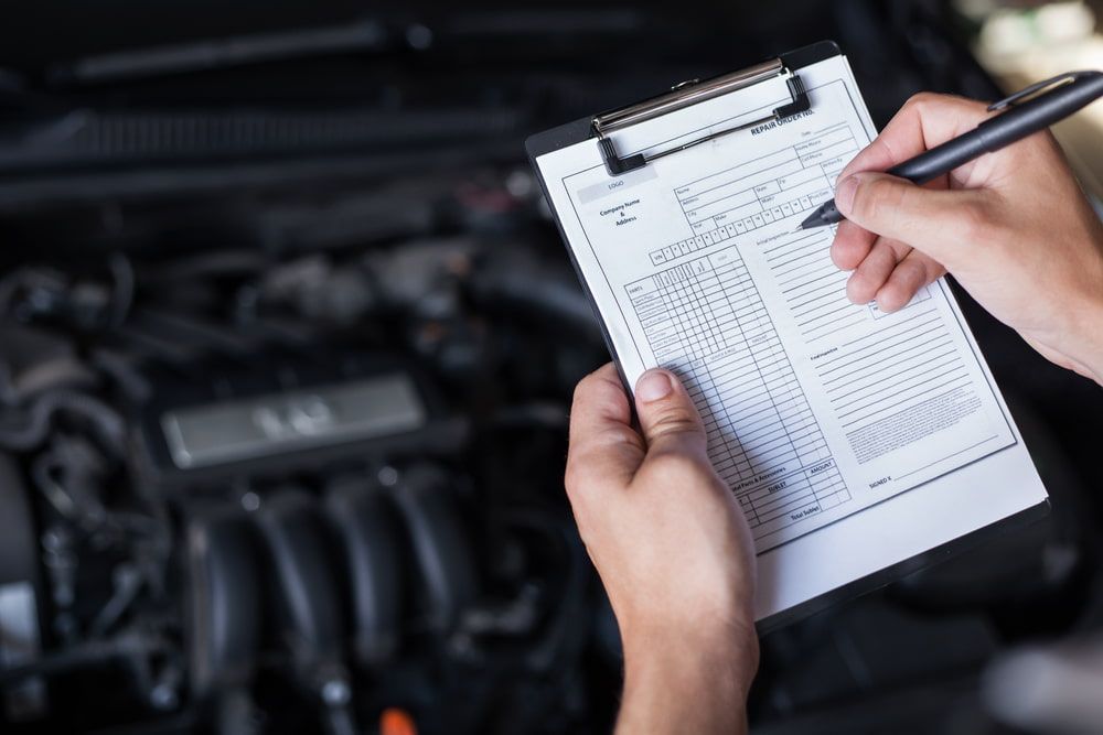 A Person Is Writing on A Clipboard in Front of A Car Engine — Allan Ross Mechanical Repairs in Charmhaven, NSW