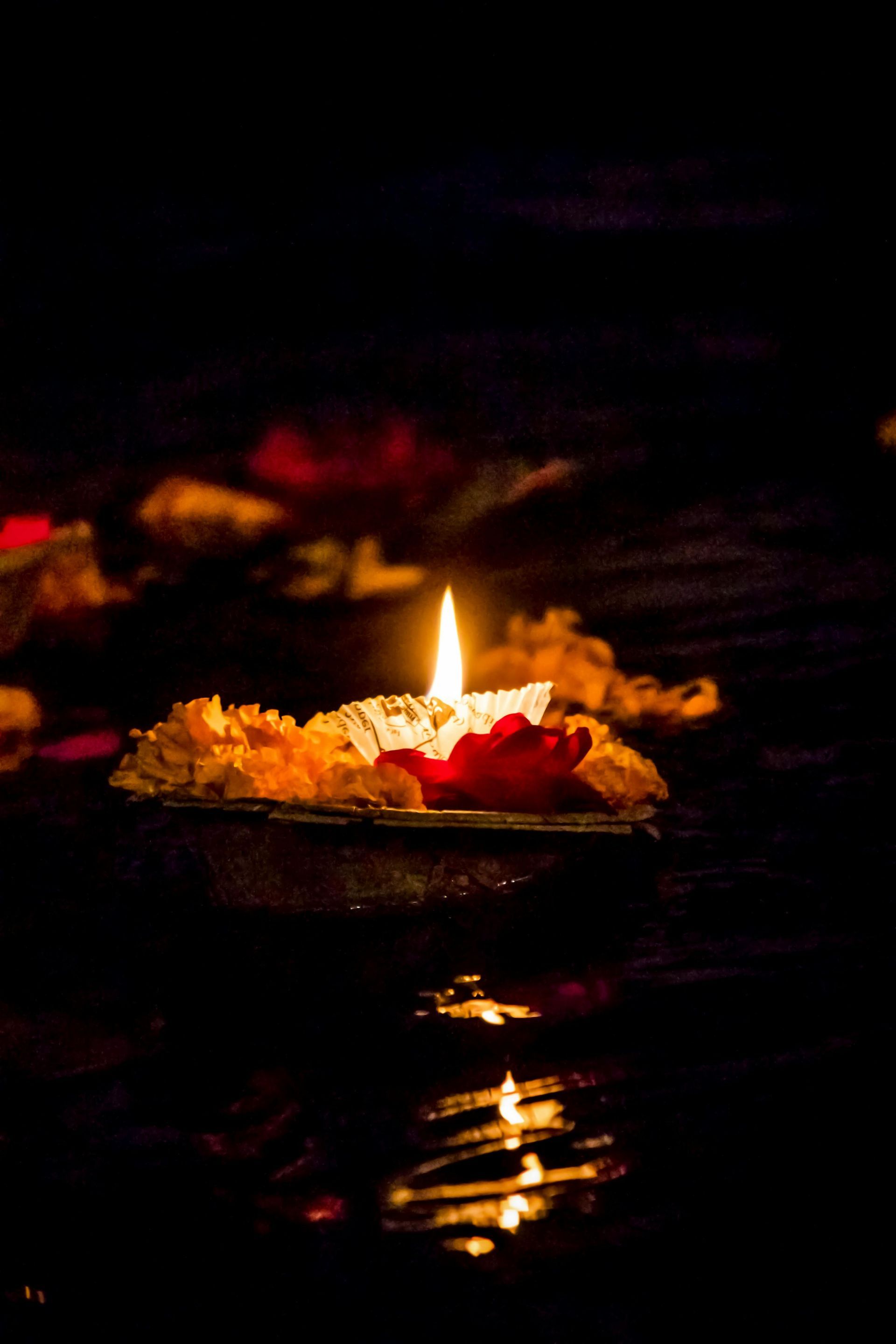 Lit candle floating on water, surrounded by flowers, against a dark background.