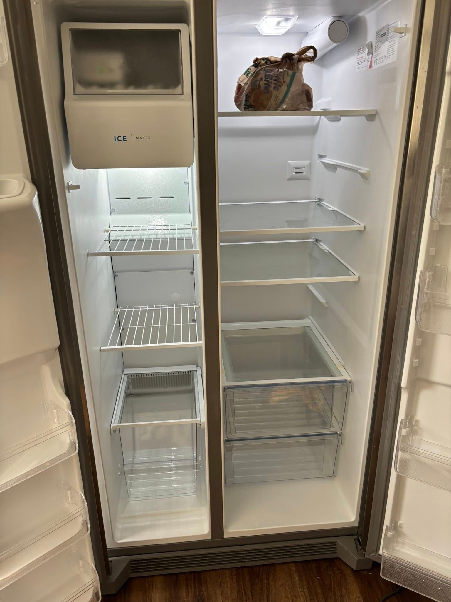 Open, empty side-by-side refrigerator, with ice maker in the freezer door, and one bag on the top shelf.