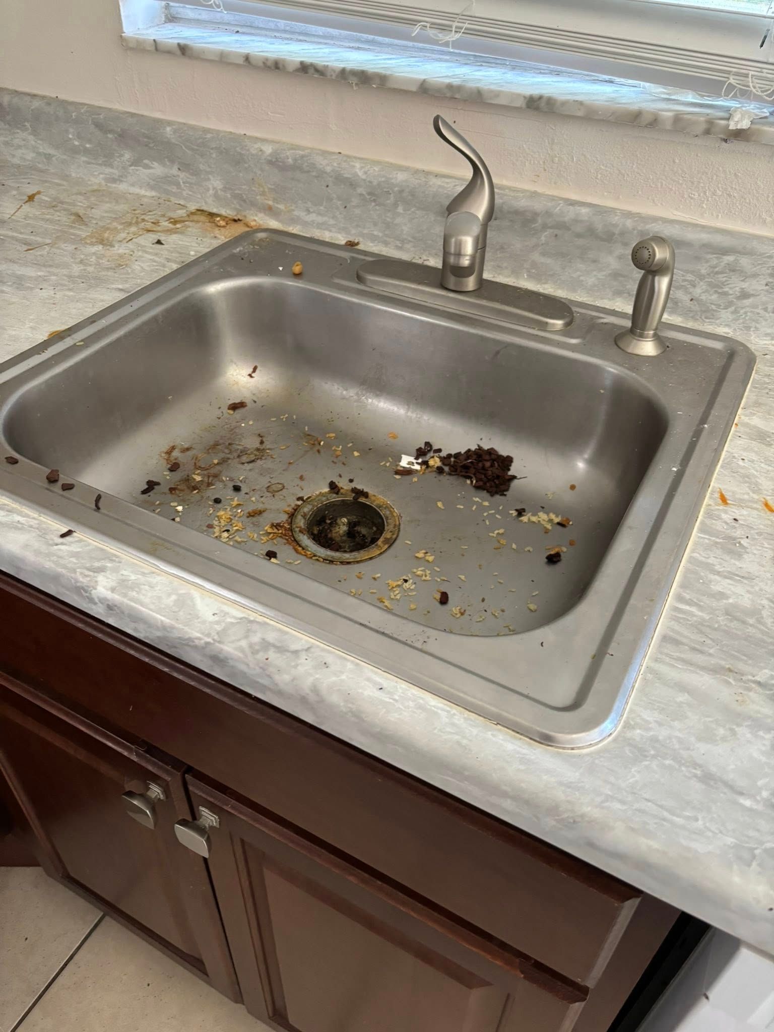 A dirty stainless steel kitchen sink filled with food scraps, faucet, and soap dispenser.