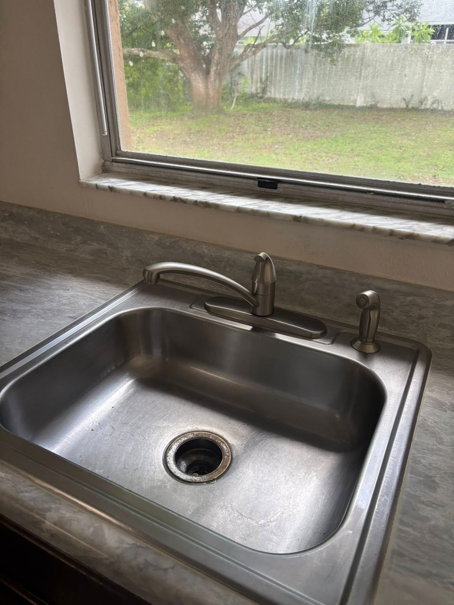 Stainless steel kitchen sink with faucet, soap dispenser, and window view of a backyard.