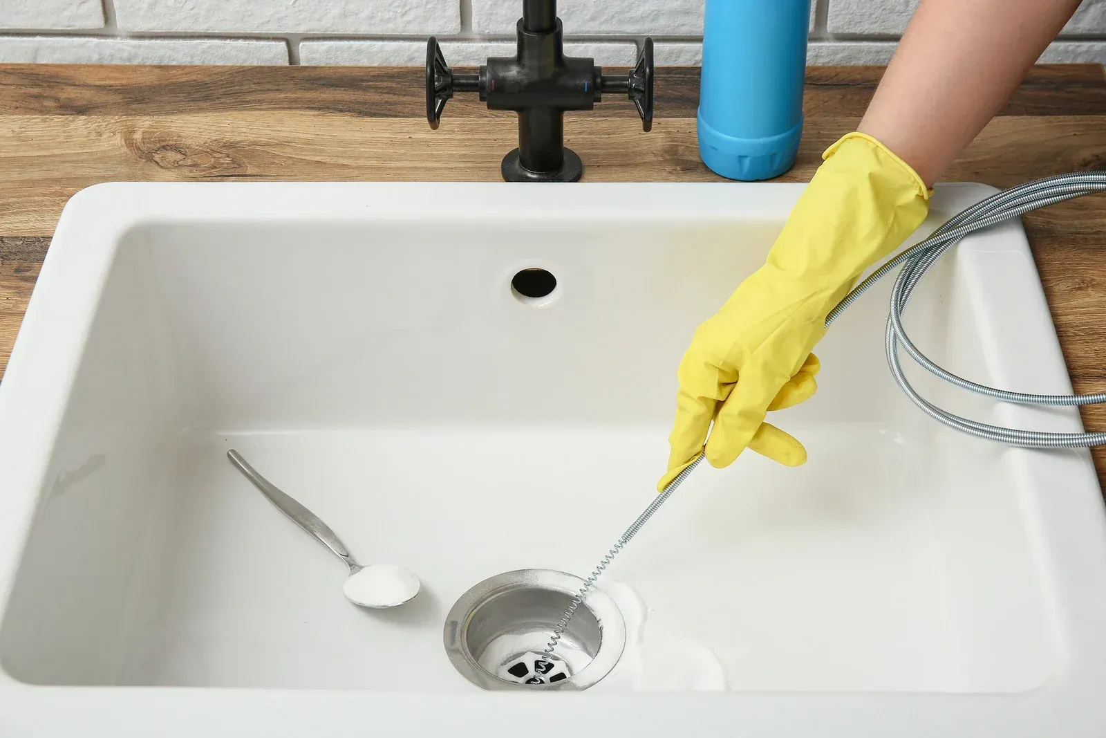 Gloved hand cleaning a white kitchen sink drain with a flexible brush, spoon nearby