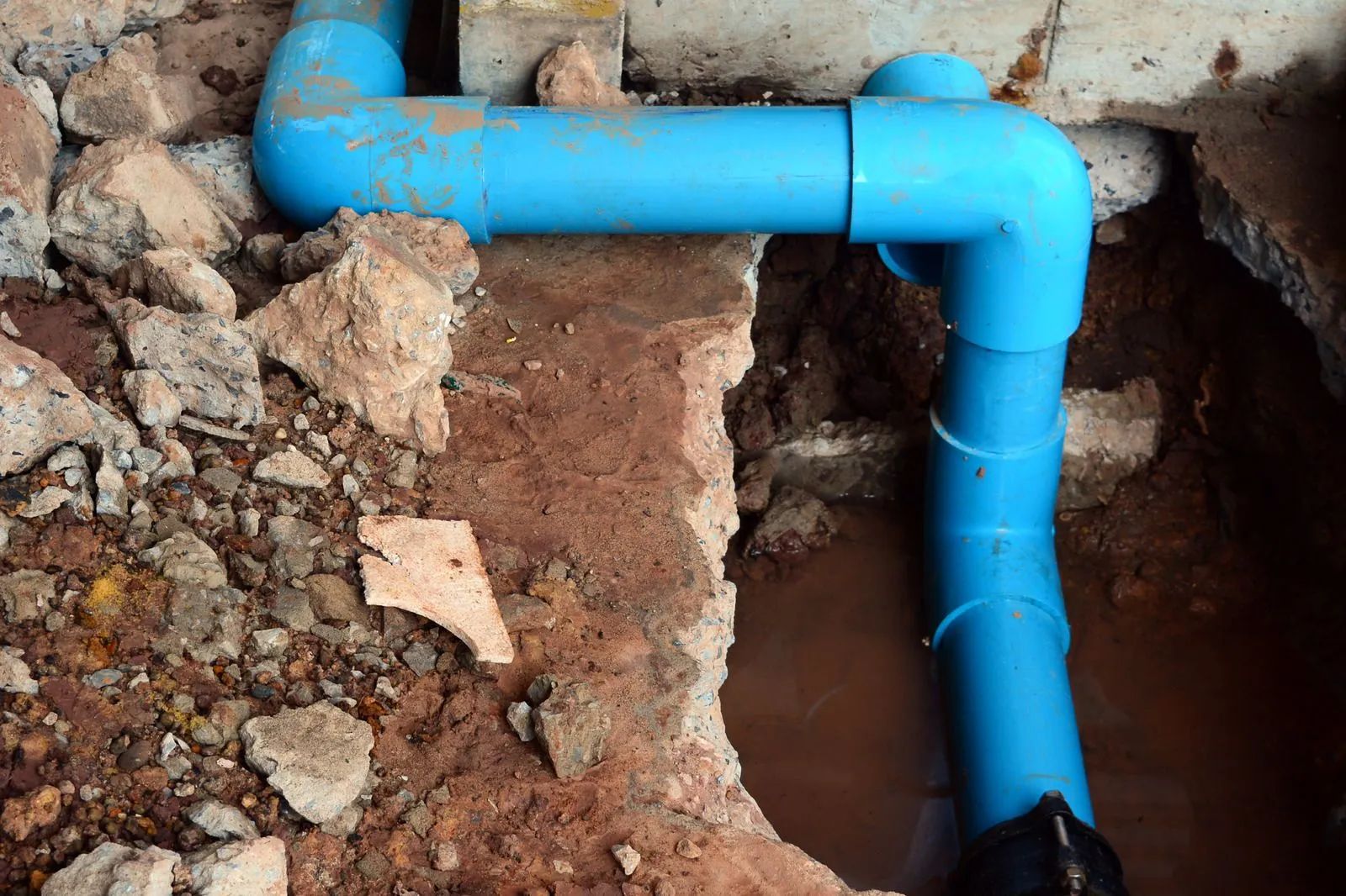 Bright blue PVC pipe running through a rough trench in broken soil and concrete rubble