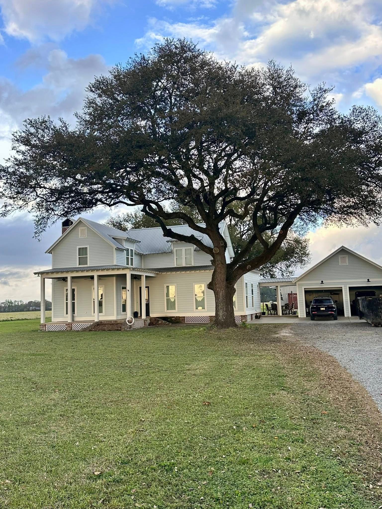 Yellow farmhouse with a large oak tree and detached garage beside a gravel drive under a cloudy sky