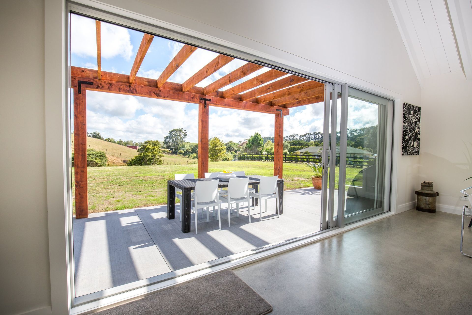 A modern, open-plan room with sliding glass doors opening to a covered patio featuring a dining table and wooden pergola.