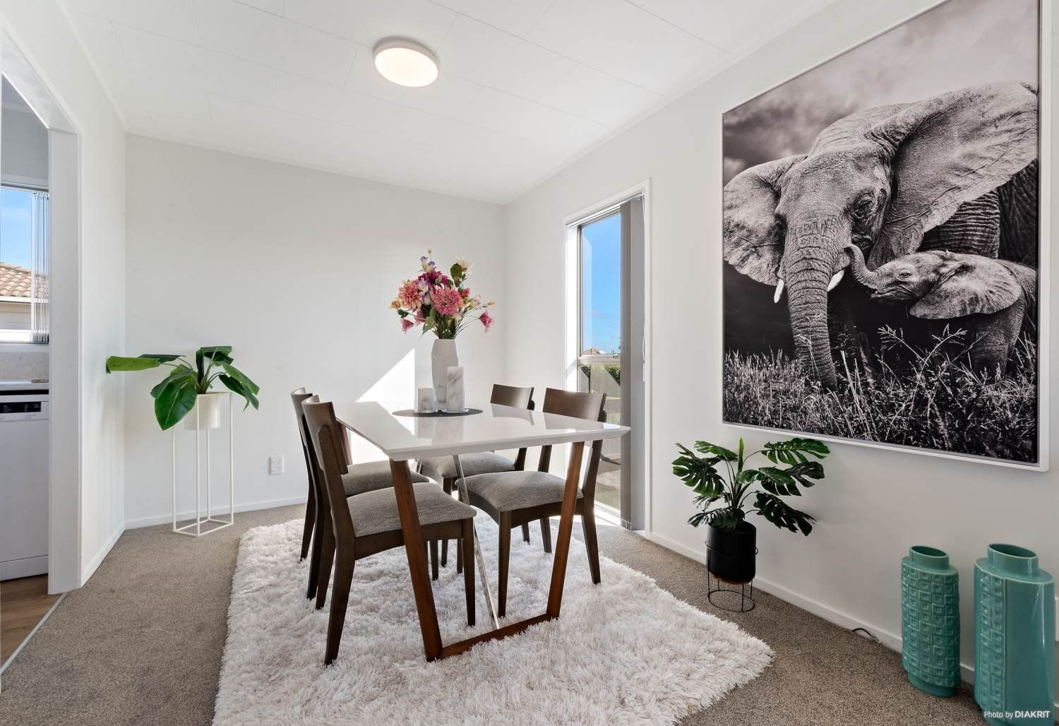 A bright dining area featuring a white table, four chairs on a fuzzy rug, a large elephant print, and decorative plants.