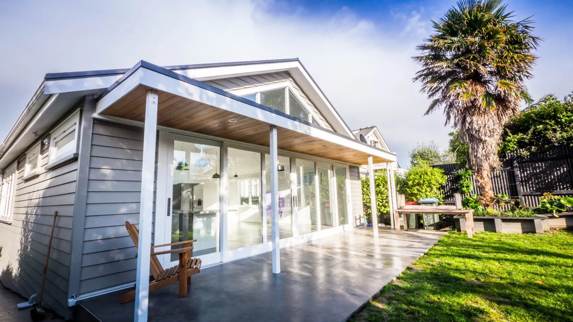A grey, modern single-story house with a large glass front, a covered patio, and a grassy yard under a bright, sunny sky.