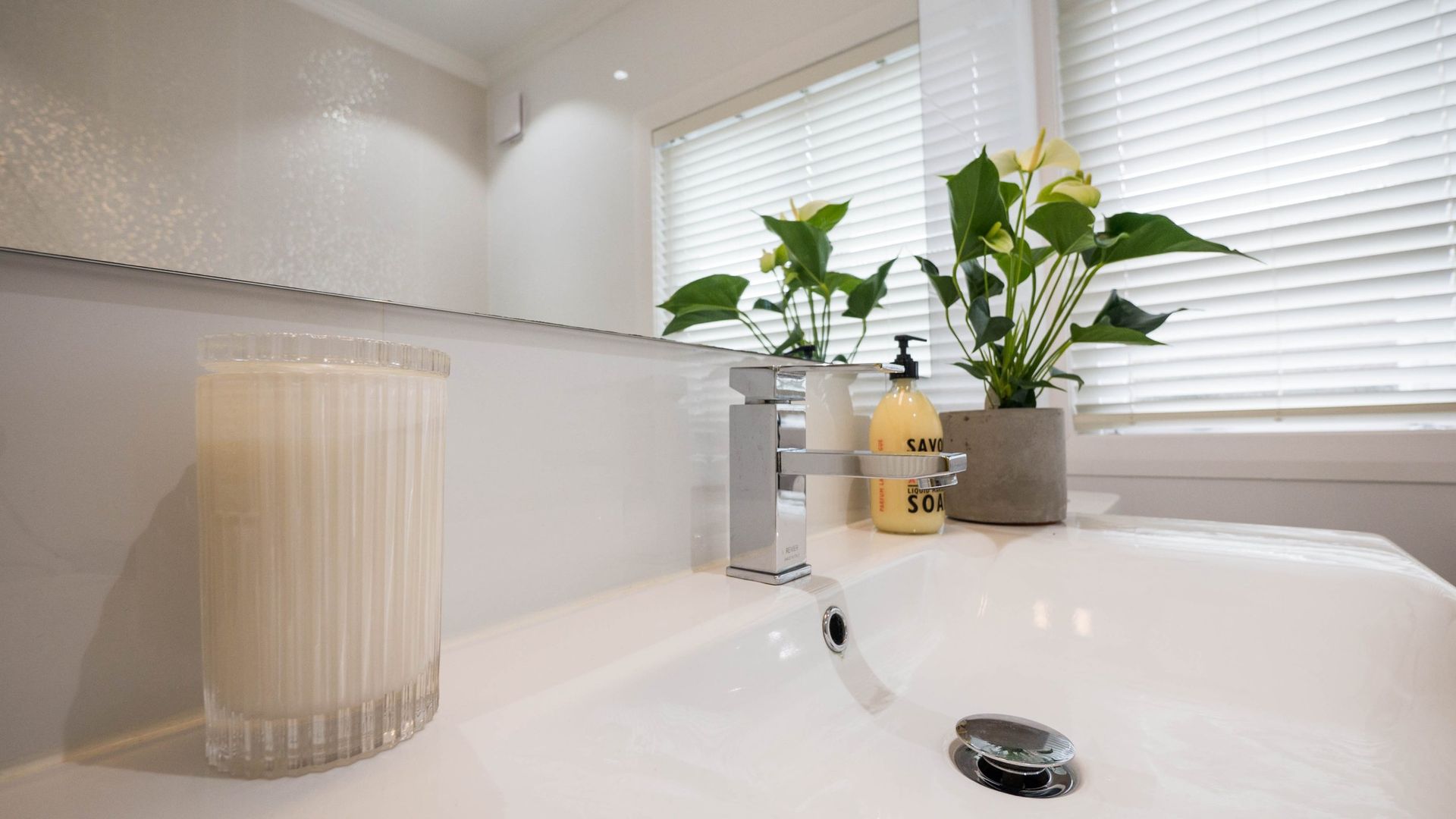 A clean white bathroom vanity with a candle, a modern faucet, and a green plant in a pot, reflected in a large mirror.
