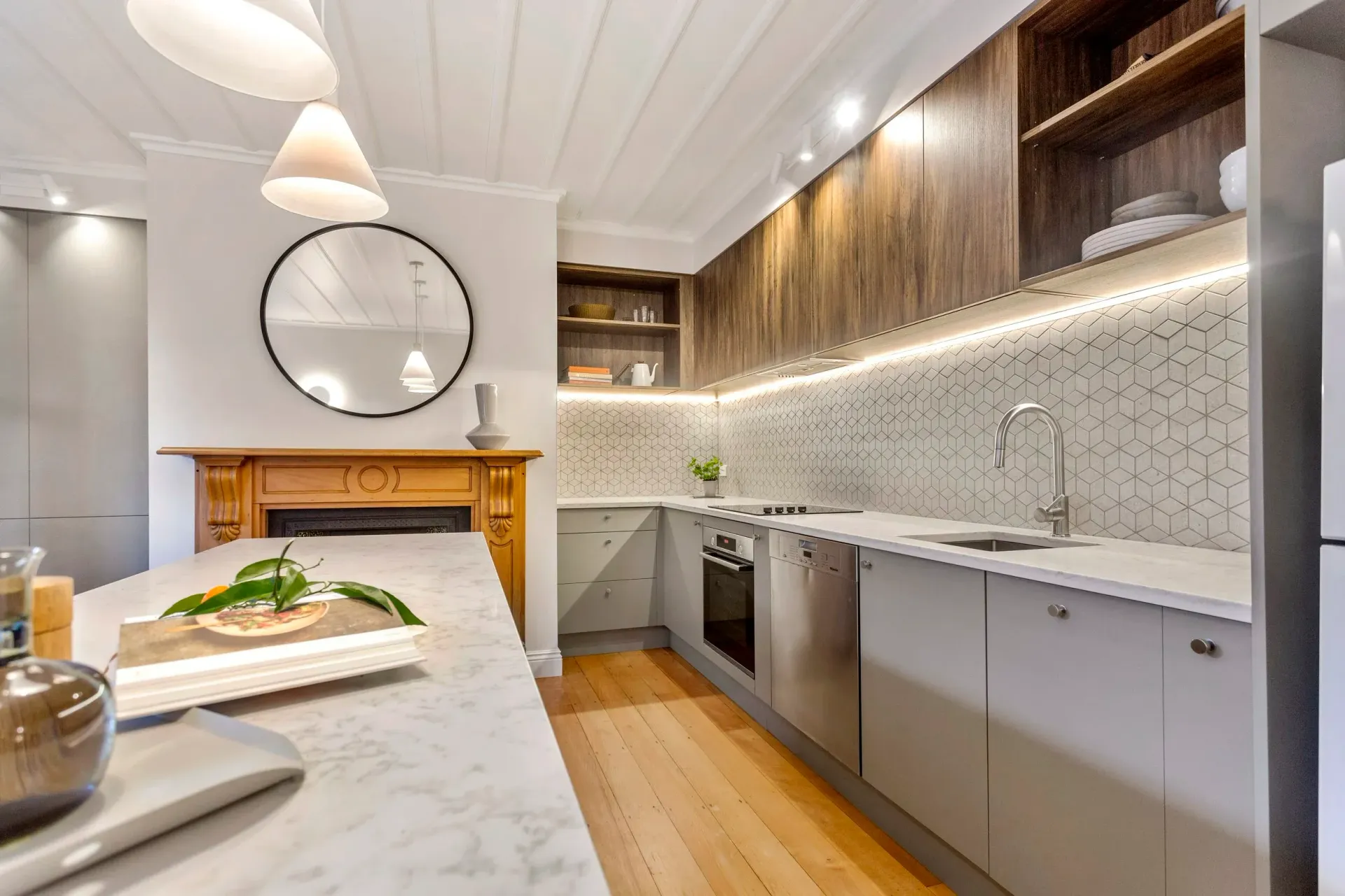 A modern kitchen with marble island, wooden fireplace, grey cabinets, patterned backsplash, and warm pendant lighting.