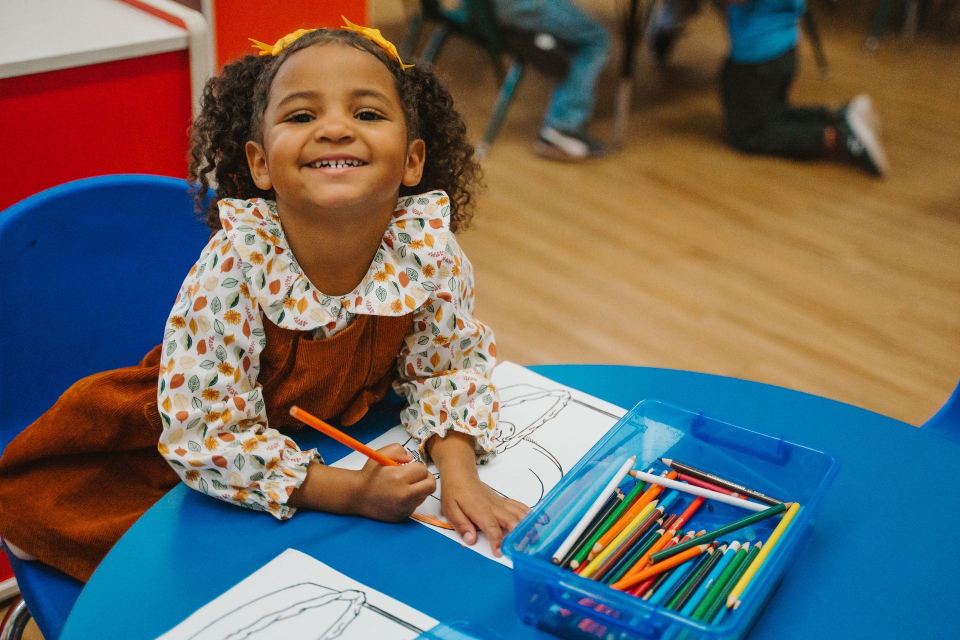 A little girl is sitting at a table with a box of colored pencils