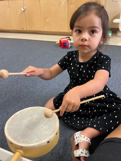 A little girl is playing a drum with wooden sticks
